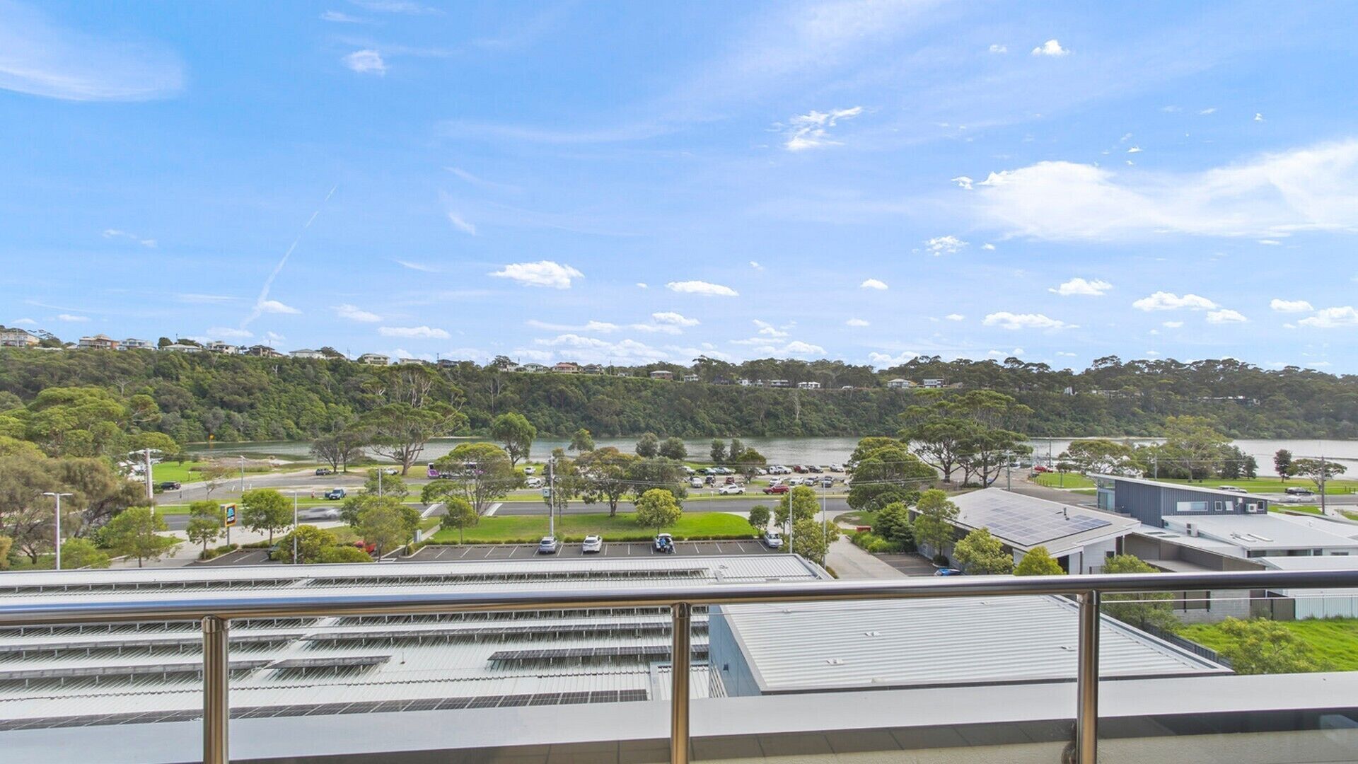 Une vue d’un parking avec plusieurs voitures, d’un bâtiment doté d’un toit plat et d’un paysage vallonné, avec des arbres et des maisons en arrière-plan.