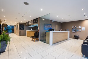 A modern reception area with a glass partition, a wooden reception desk, and a seating area with chairs. - The Esplanade Resort and Spa (Lakes Entrance)