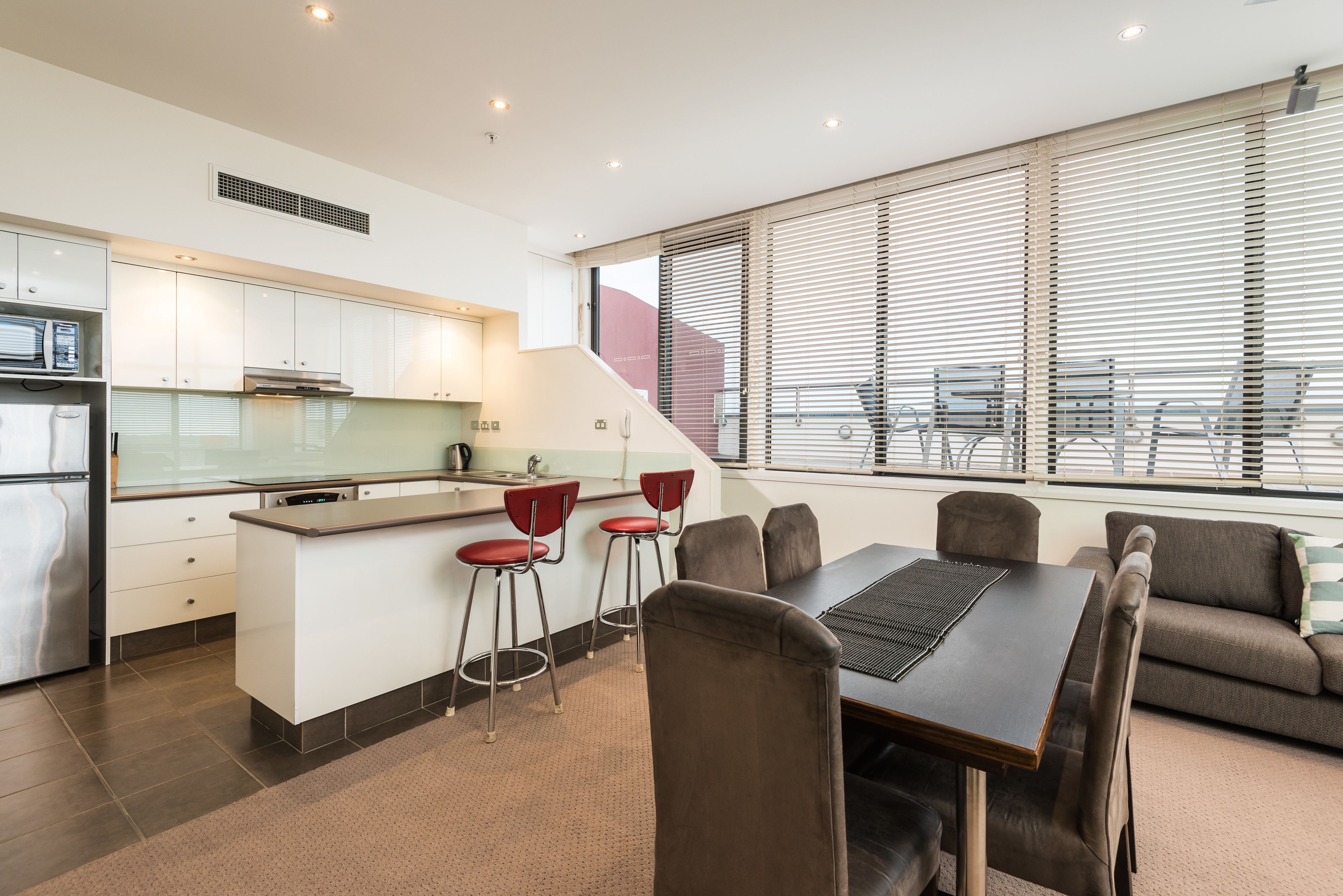 A modern kitchen with white cabinets, a central island, and red bar stools. There is a dining area with a table and chairs, and a sofa.