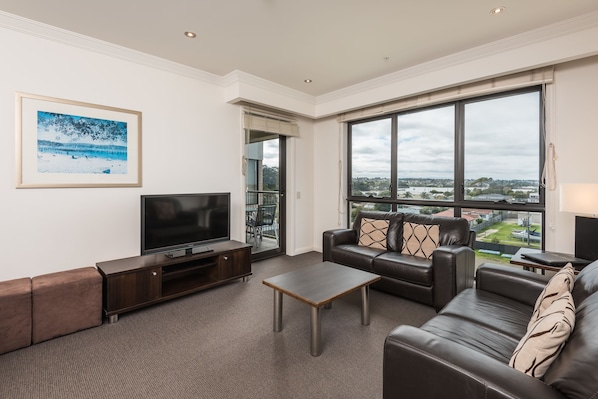 A modern living room with a large window, a flat-screen TV, and a leather sofa. - The Esplanade Resort and Spa (Lakes Entrance)