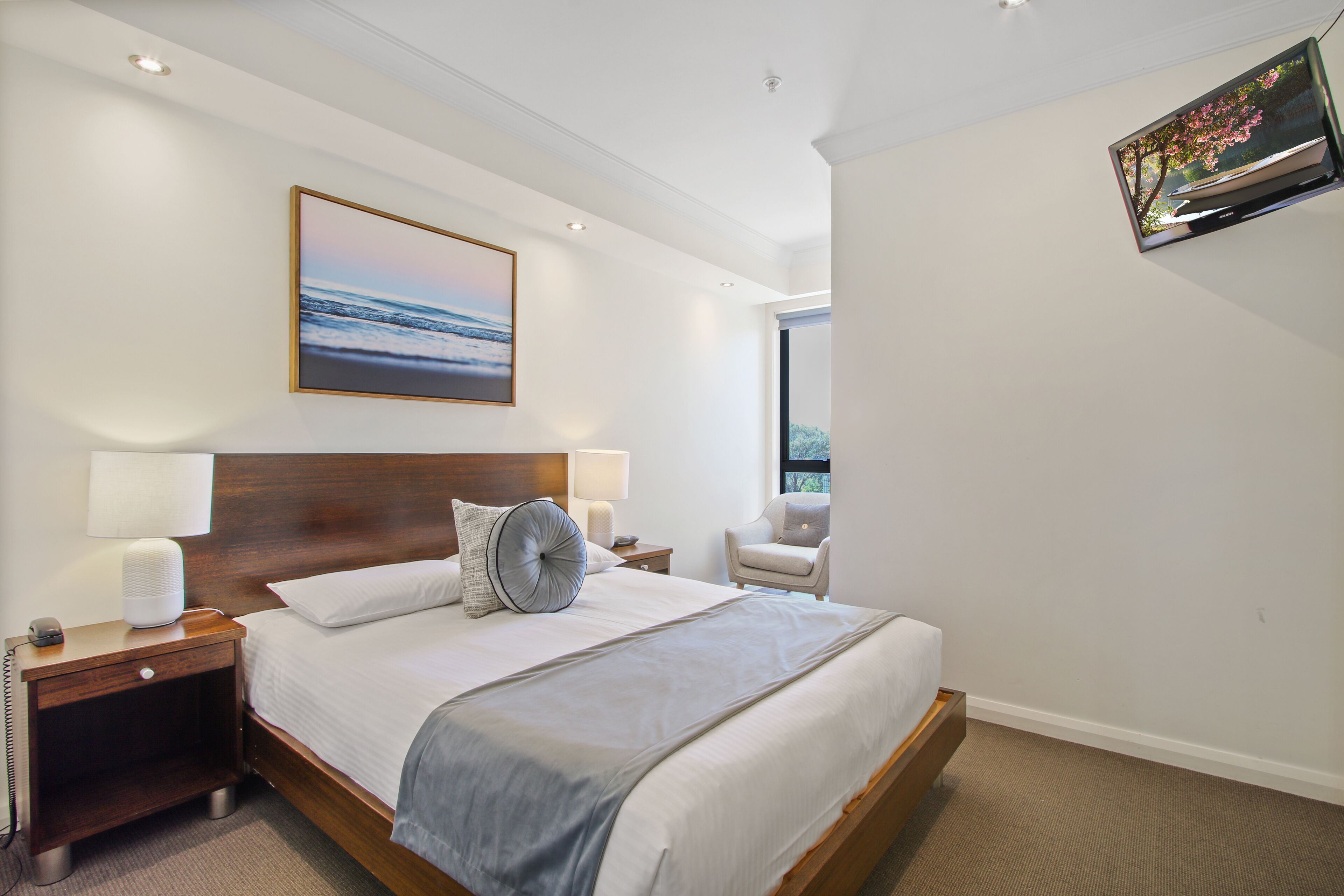 Una habitación de hotel moderna con una cama grande, lámparas de noche, un televisor montado y un cuadro con una escena de playa.