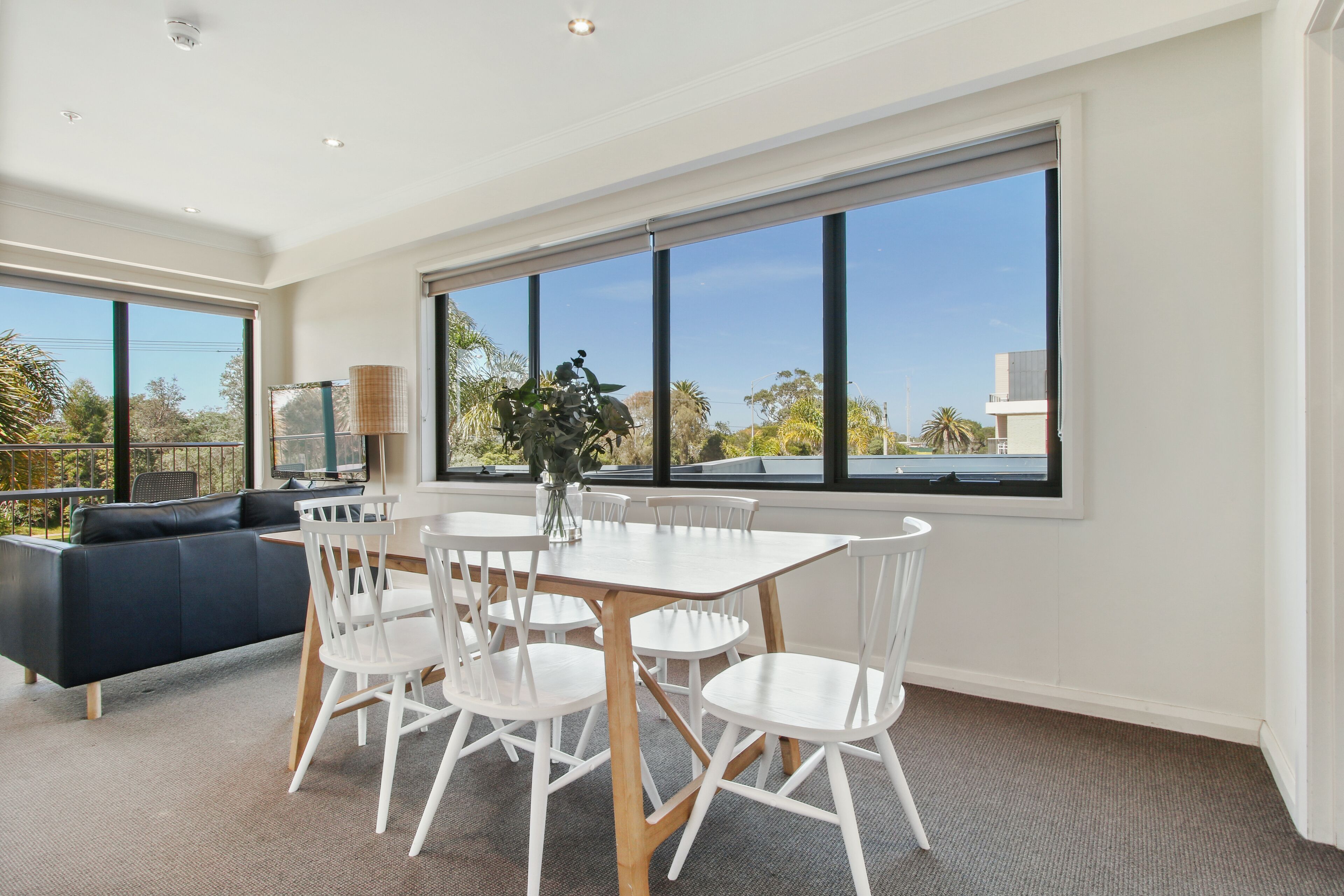 A modern dining area with a white table and chairs, a black sofa, and large windows offering a view of the outdoors.