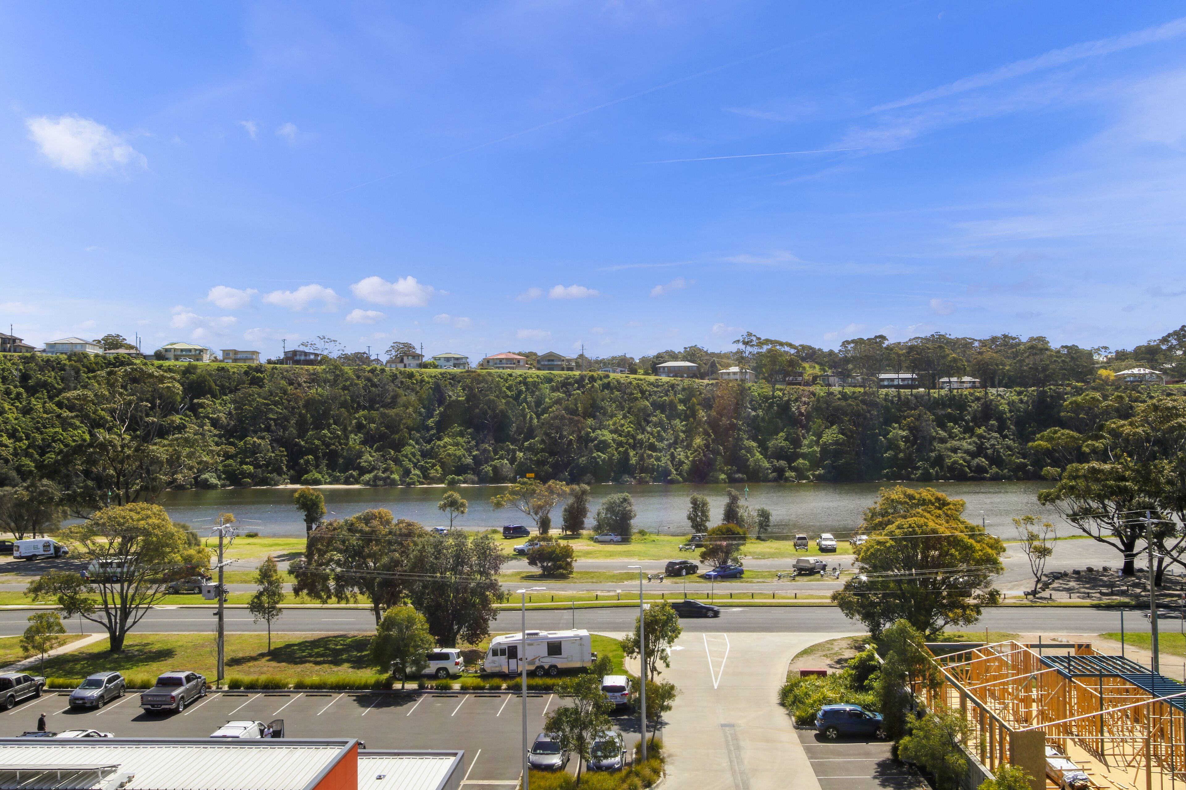 A parking lot with several cars and a caravan, surrounded by greenery and a lake.