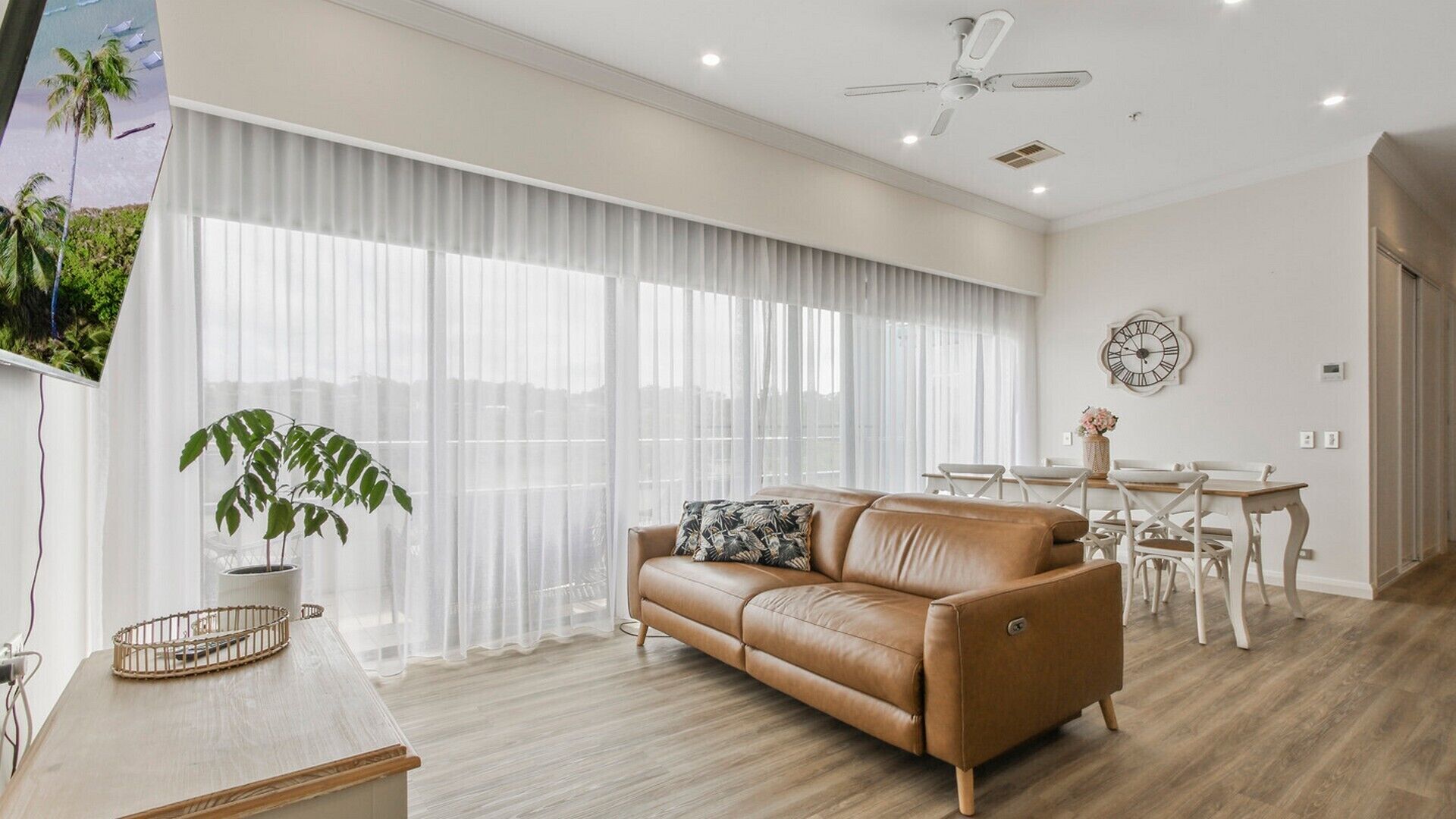 A modern living room with a leather sofa, a dining table with chairs, and a large window with sheer curtains.