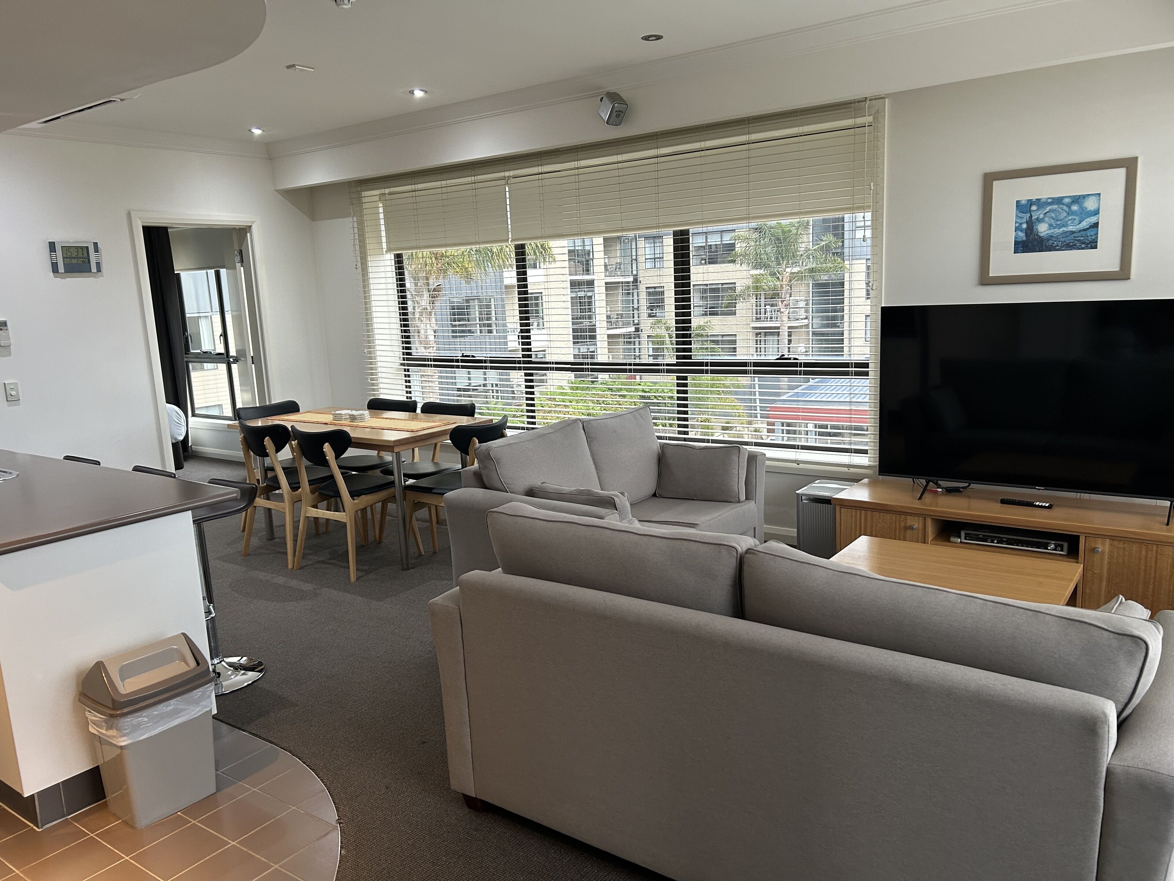 Ein modernes Wohnzimmer mit einem grauen Sofa, einem Esstisch mit Stühlen, einem Fernseher und einem Fenster mit Jalousien.