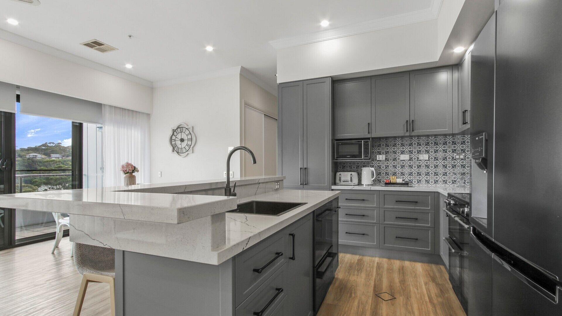 A modern kitchen with grey cabinets, a large island with a sink, a microwave, and a refrigerator.
