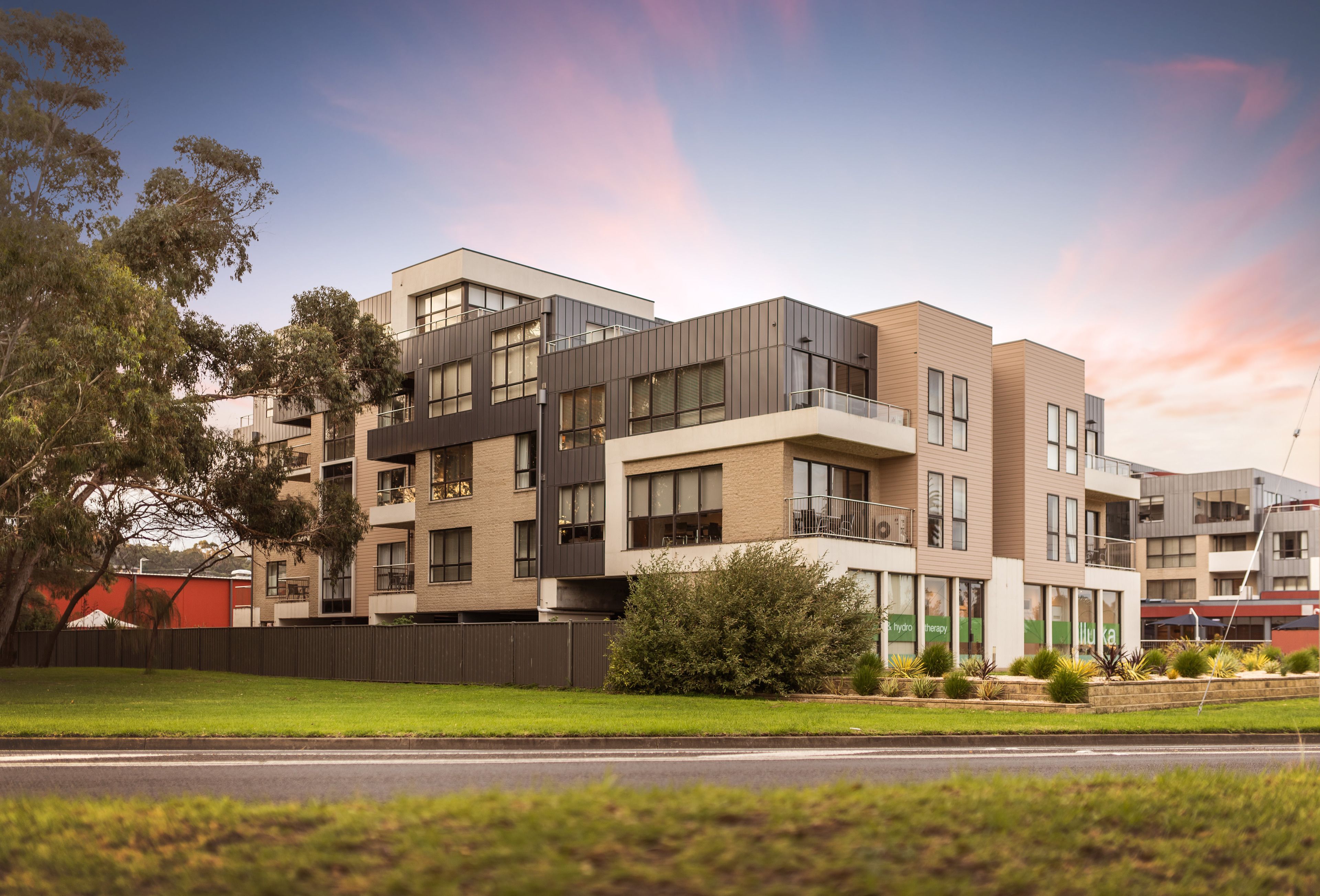 A modern multi-story residential building with balconies, large windows, and a landscaped front yard.