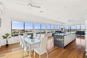 A spacious modern kitchen with a large island, large windows with a view of the water, and a dining area with white chairs and a glass table. - The Esplanade Resort and Spa (Lakes Entrance)