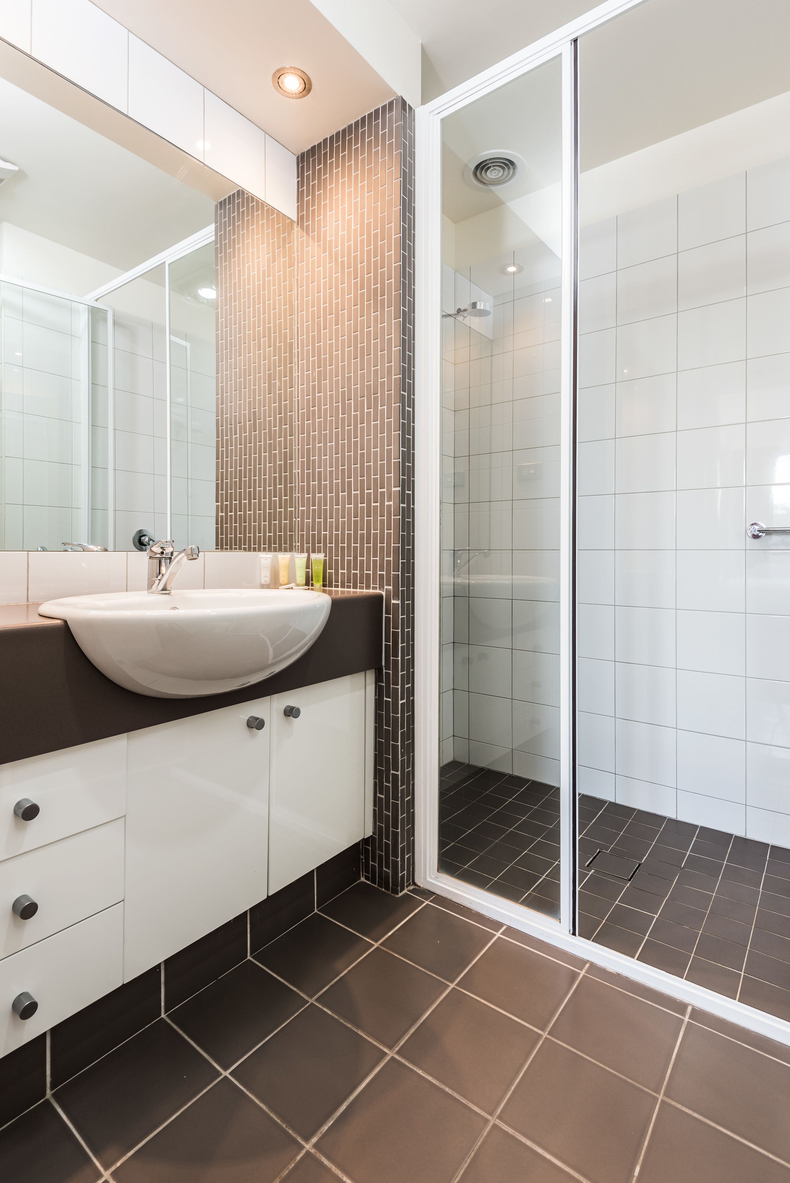 Une salle de bain moderne avec un lavabo blanc, des carreaux de couleur marron foncé et une cabine de douche en verre.