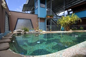 Una piscina con borde de piedra, cercada por una valla metálica, y una escalera que lleva a un edificio azul.