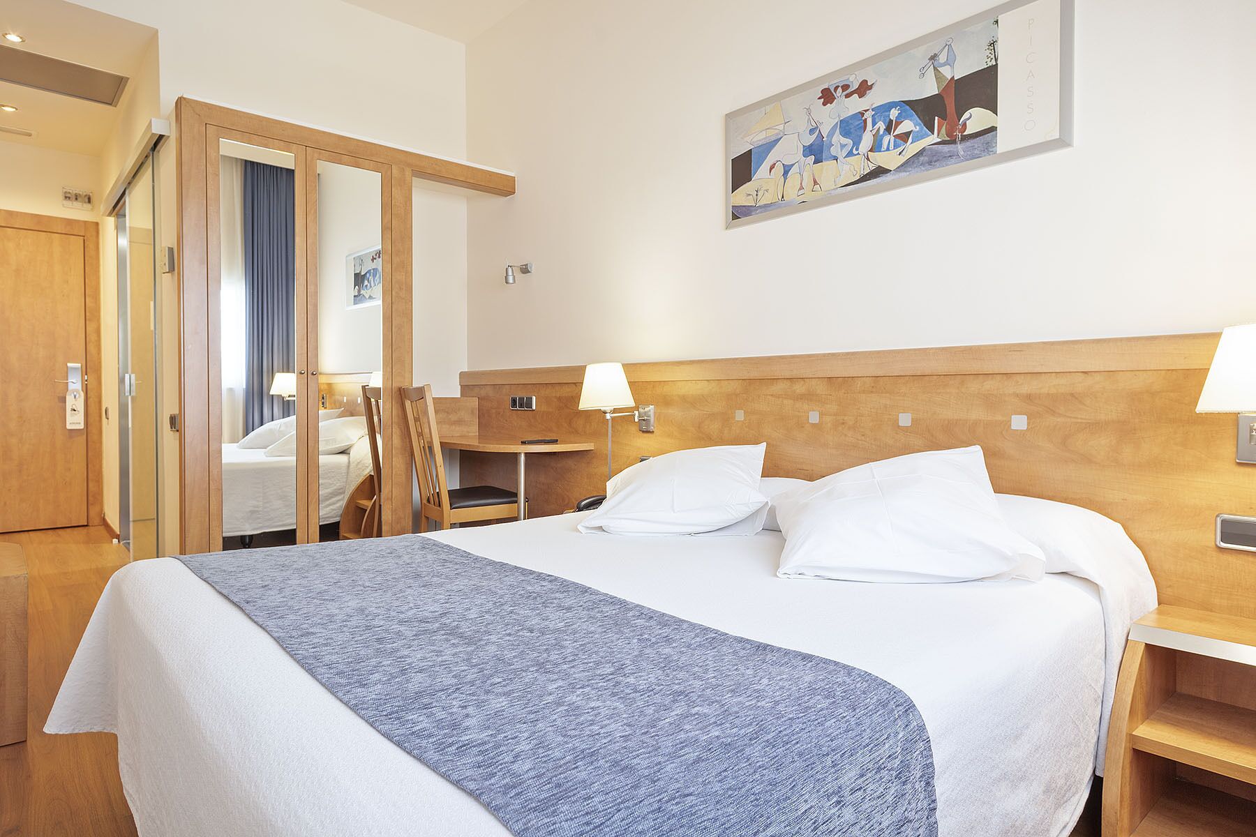 A hotel room with a large bed, wooden headboard, bedside tables with lamps, a mirror, and a painting on the wall.