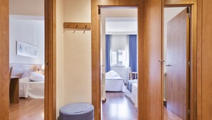 A hotel corridor with wooden doors, a small seating area, and two bedrooms with white bedding.