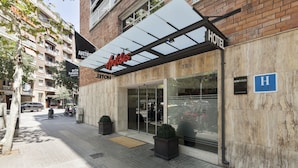 A hotel entrance with a glass facade, a covered outdoor seating area, and a clear blue sign indicating the hotel name.