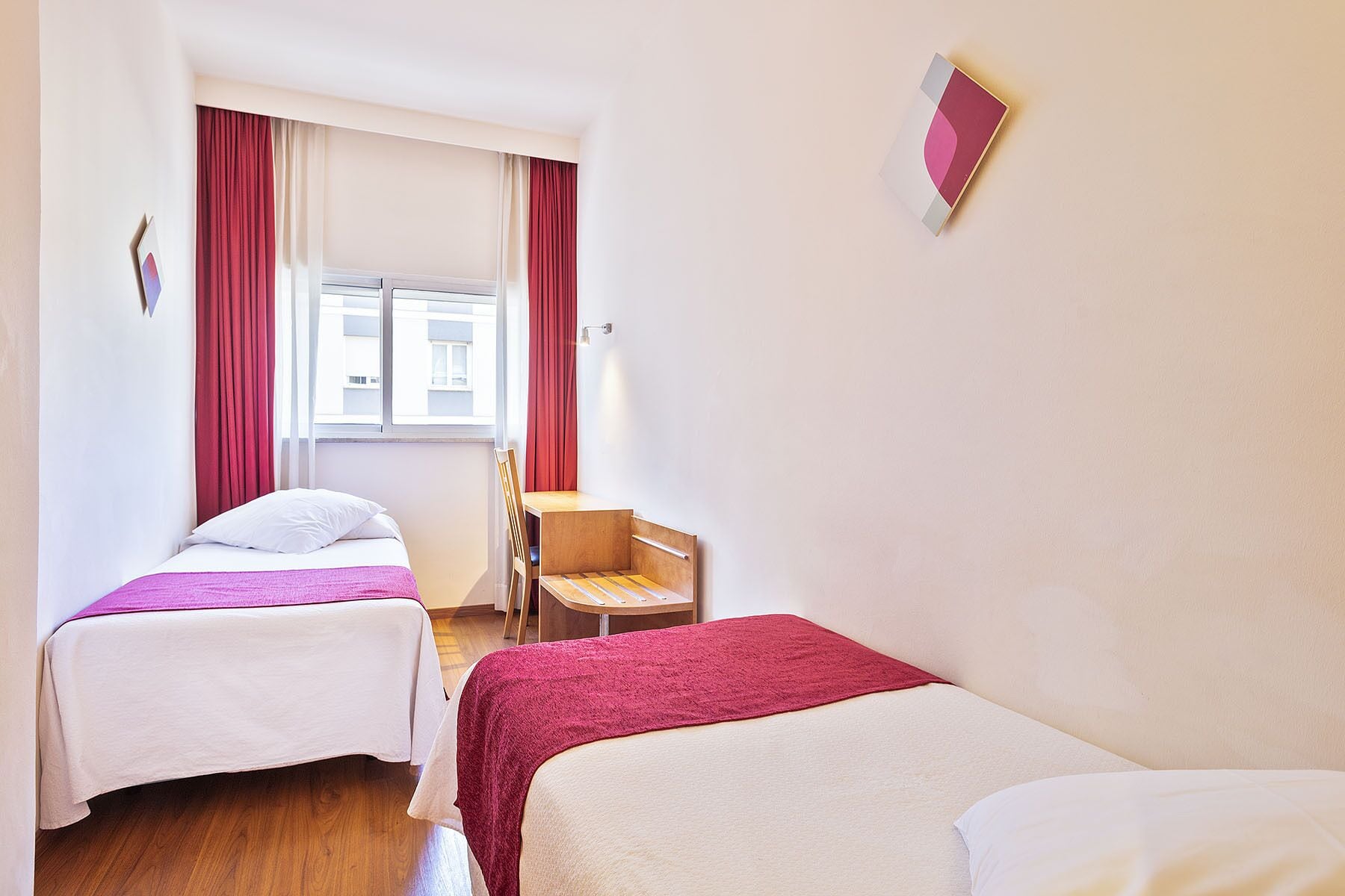 Une chambre d’hôtel avec deux lits simples, une chaise en bois, une petite table et une fenêtre avec des rideaux.