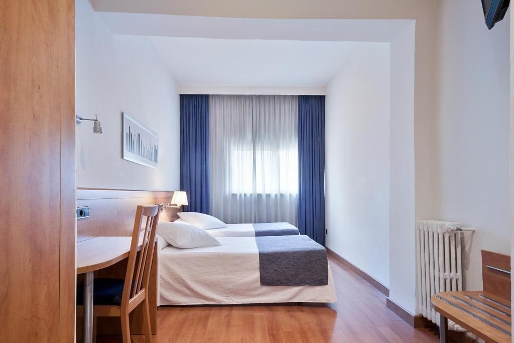 A hotel room with a wooden desk, two beds, a radiator, and a window with curtains.