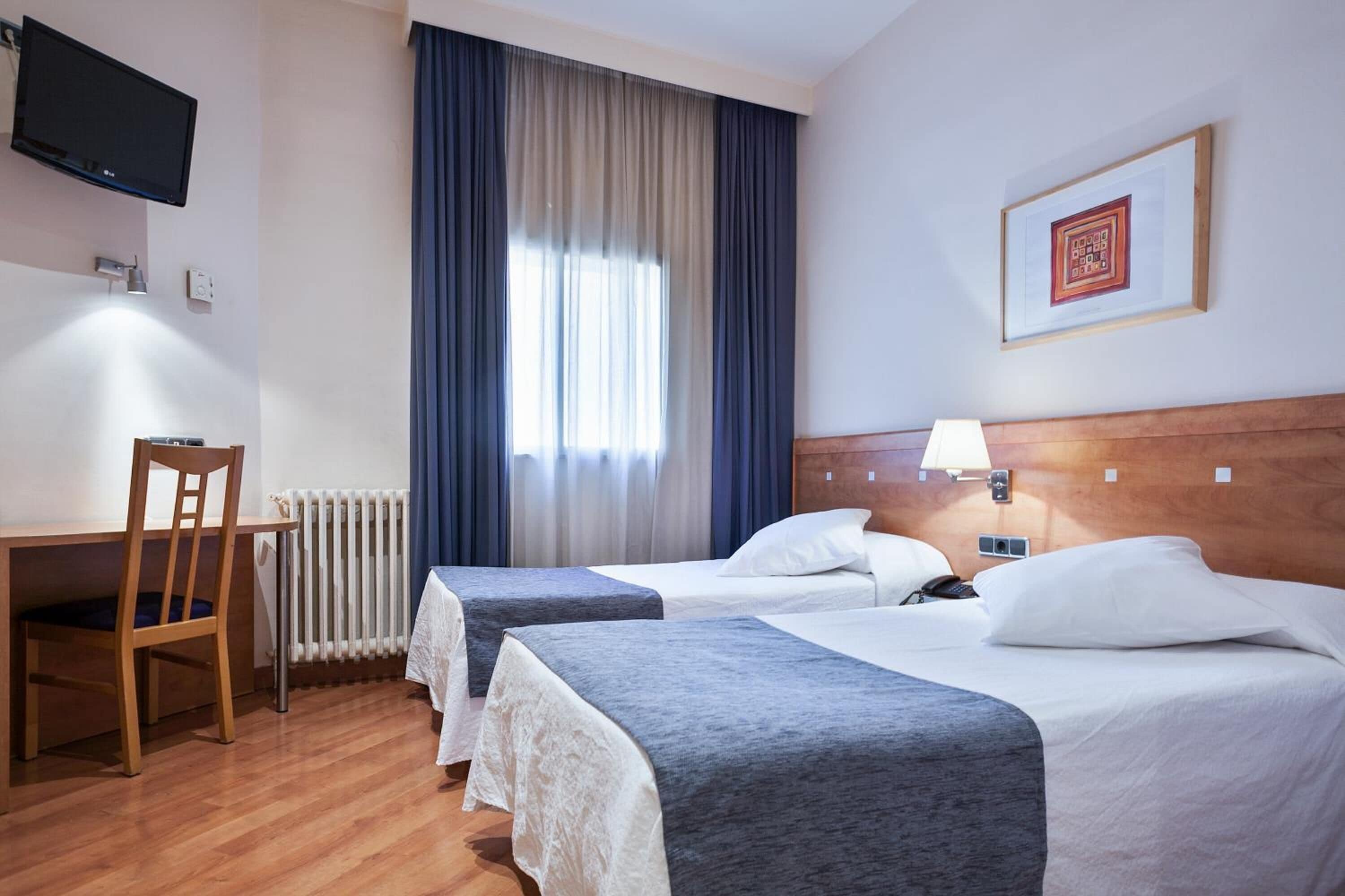A hotel room with two beds, a desk, a chair, a TV, and a window with curtains.