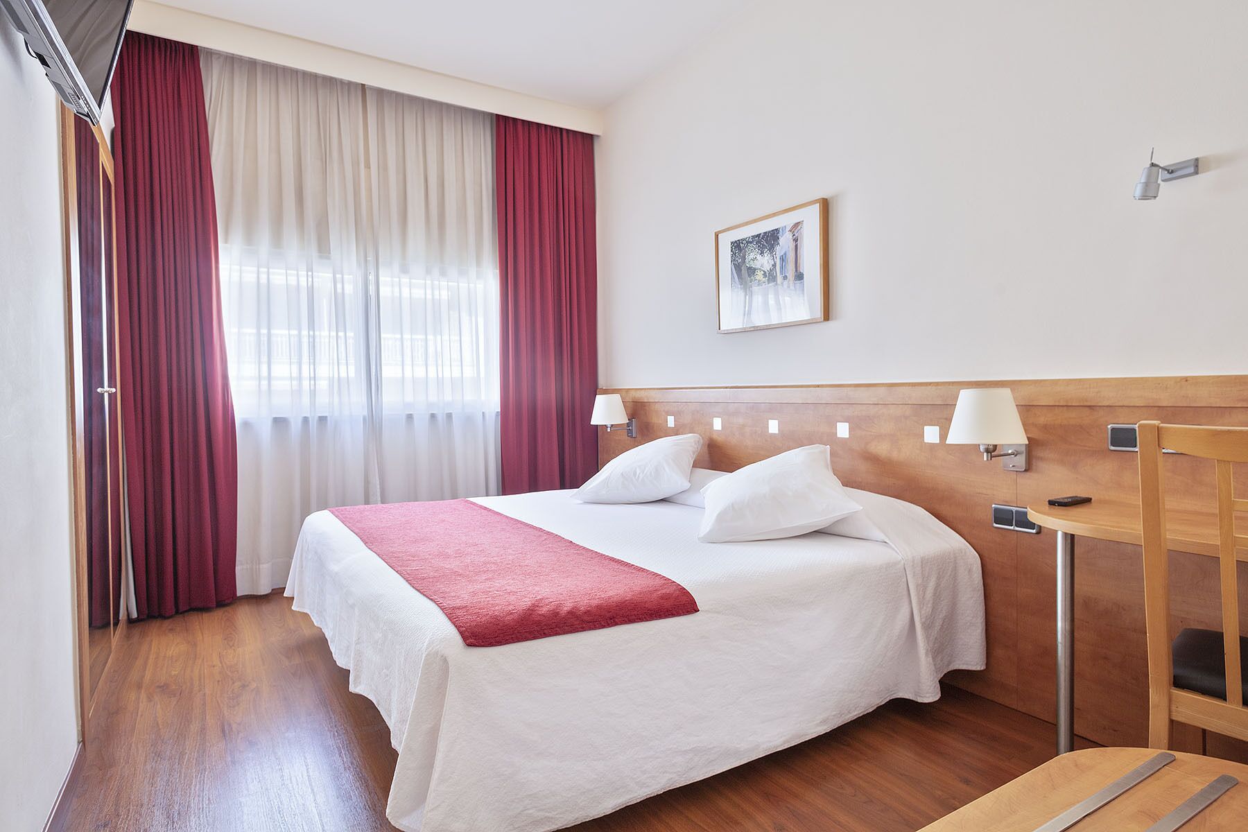 Habitación de hotel con cama, cabecera de madera, lámparas de noche, una mesita y un cuadro en la pared.