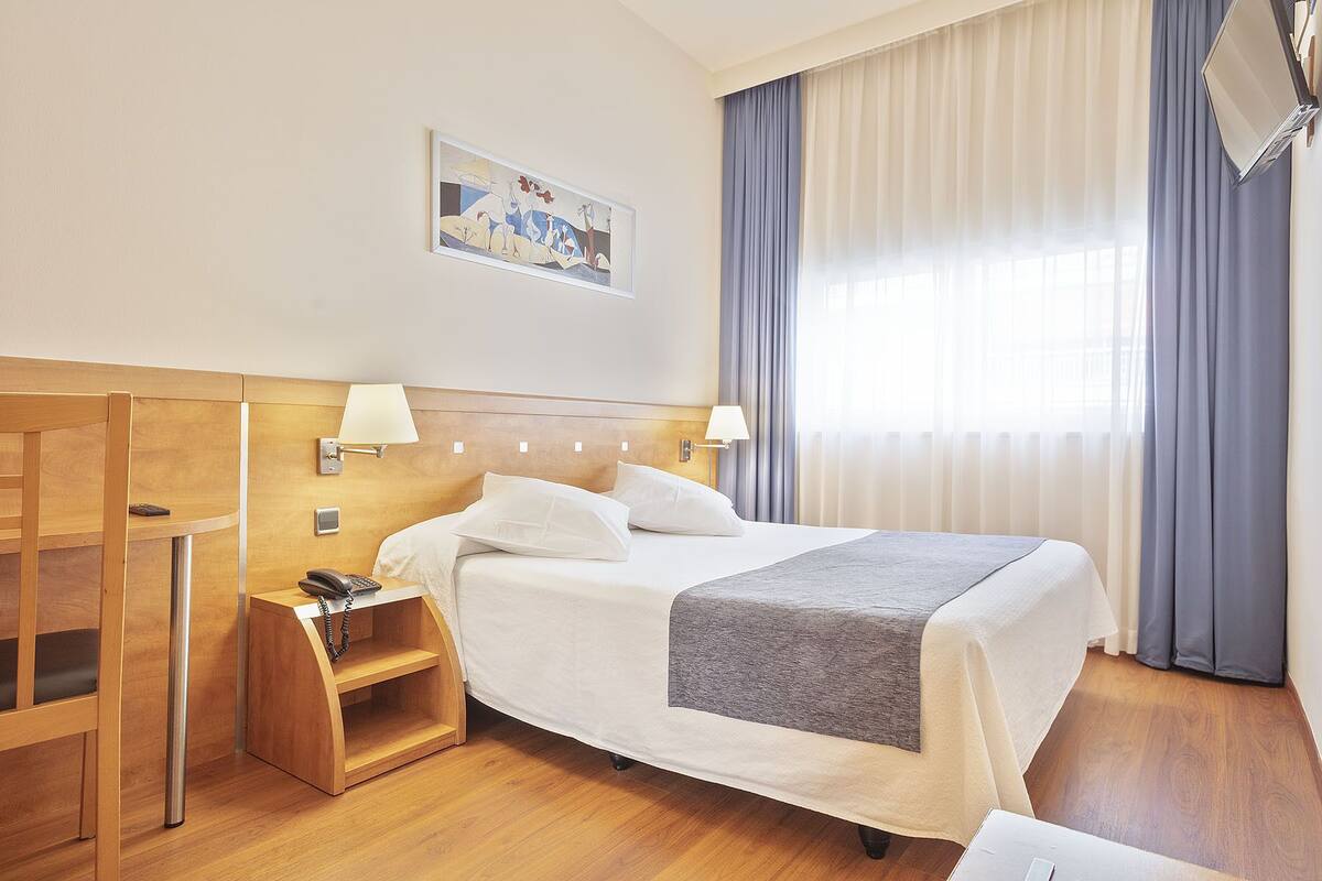 A hotel room with a bed, bedside lamps, a wooden nightstand, a telephone, and a painting on the wall.