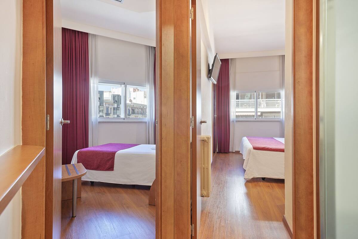 A hotel room with two beds, a wooden desk, and a view of the city through large windows.
