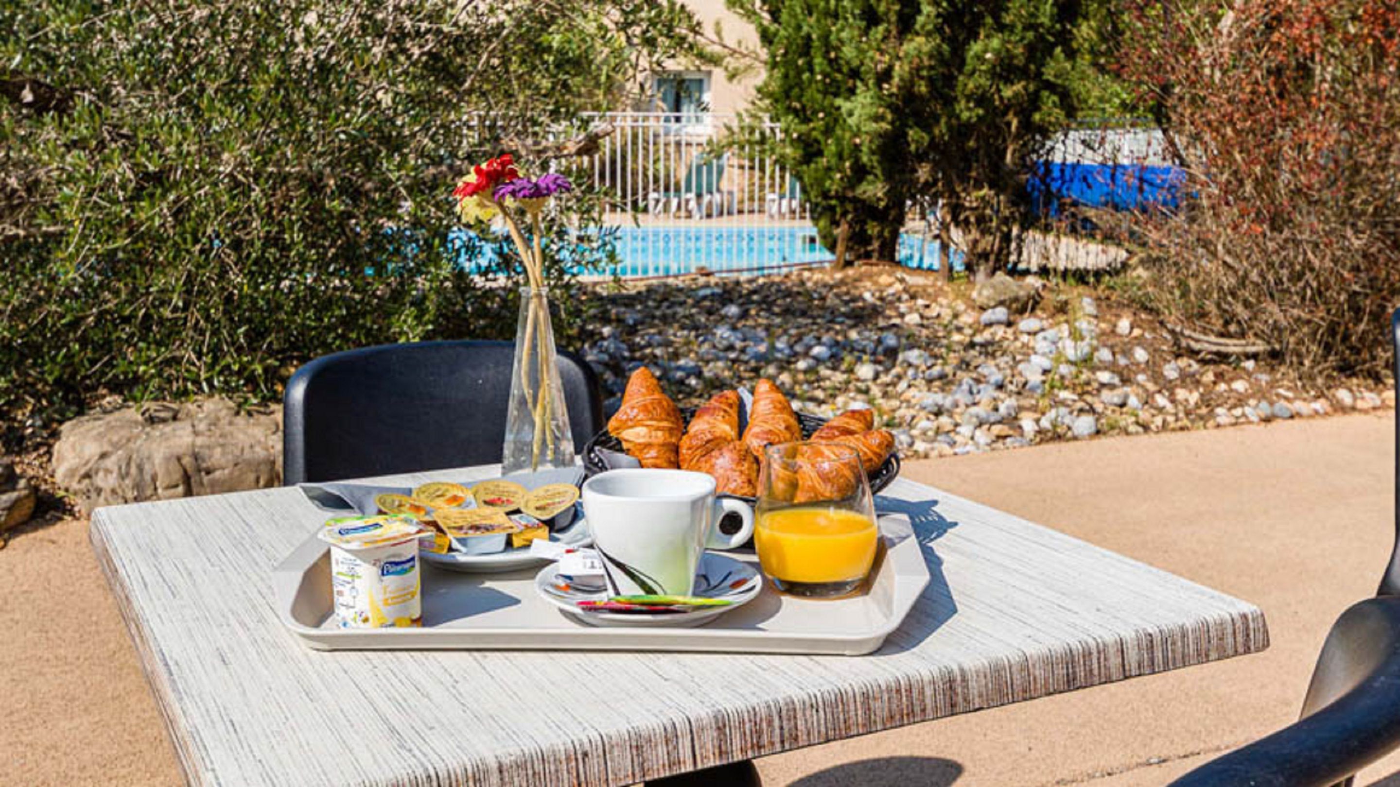 A table with breakfast items including croissants, yogurt, and orange juice, set outdoors with a pool and greenery in the background.