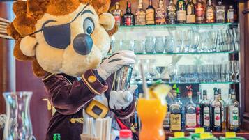 A mascot dressed in a brown and white costume with a large nose and glasses, standing in front of a bar area with various bottles and glasses.