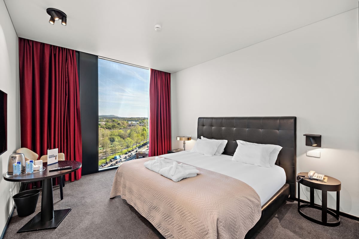 Een hotelkamer met een groot bed, een bureau met stoel, een tafel met een rood gordijn en uitzicht op een stadsbeeld.