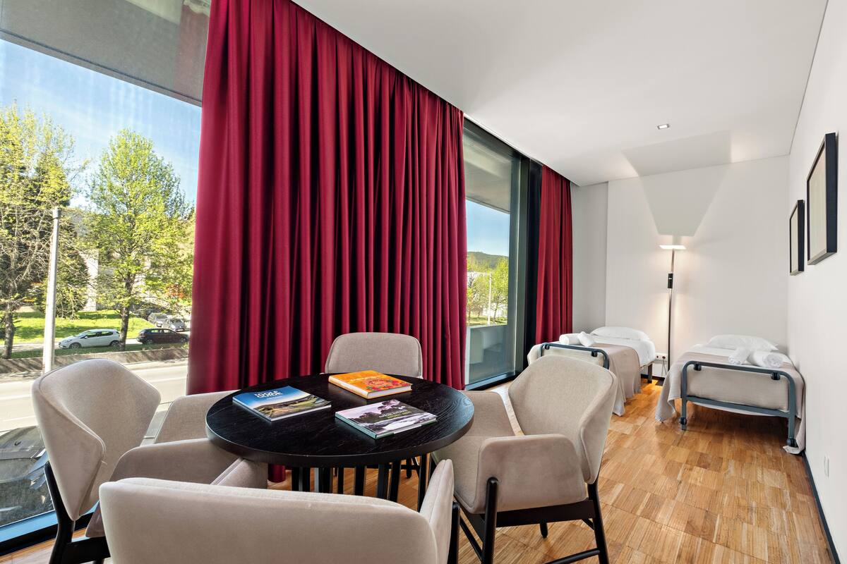 Een hotelkamer met een groot raam, een ronde tafel met stoelen, twee bedden en een lamp.