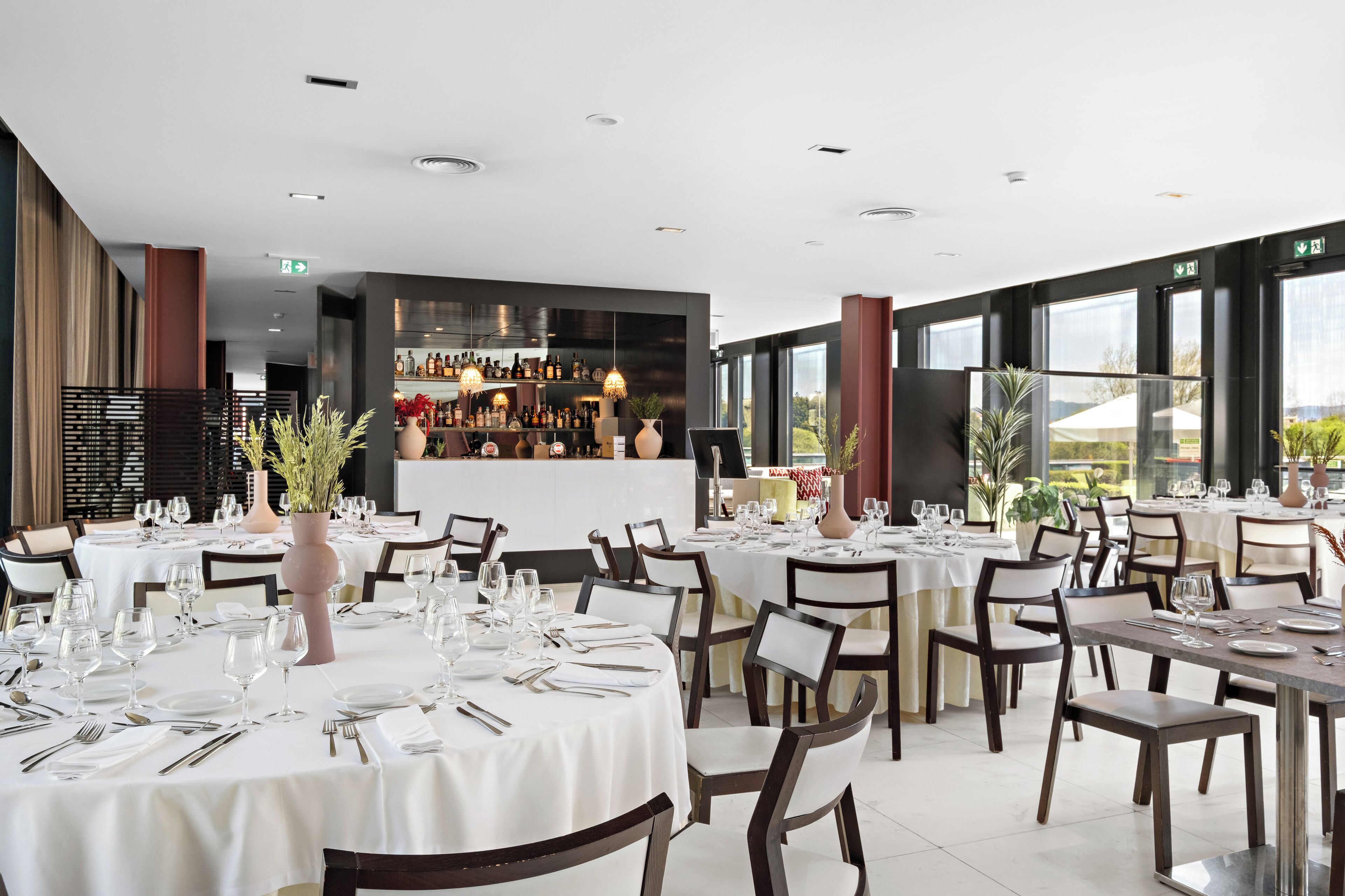 A restaurant with round tables, chairs, a bar area, and large windows.