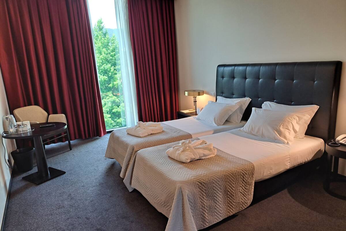 Een hotelkamer met twee bedden, een bureau, een stoel en uitzicht op groen.
