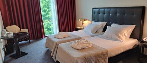 A hotel room with two beds, a desk, a chair, and a view of greenery outside.