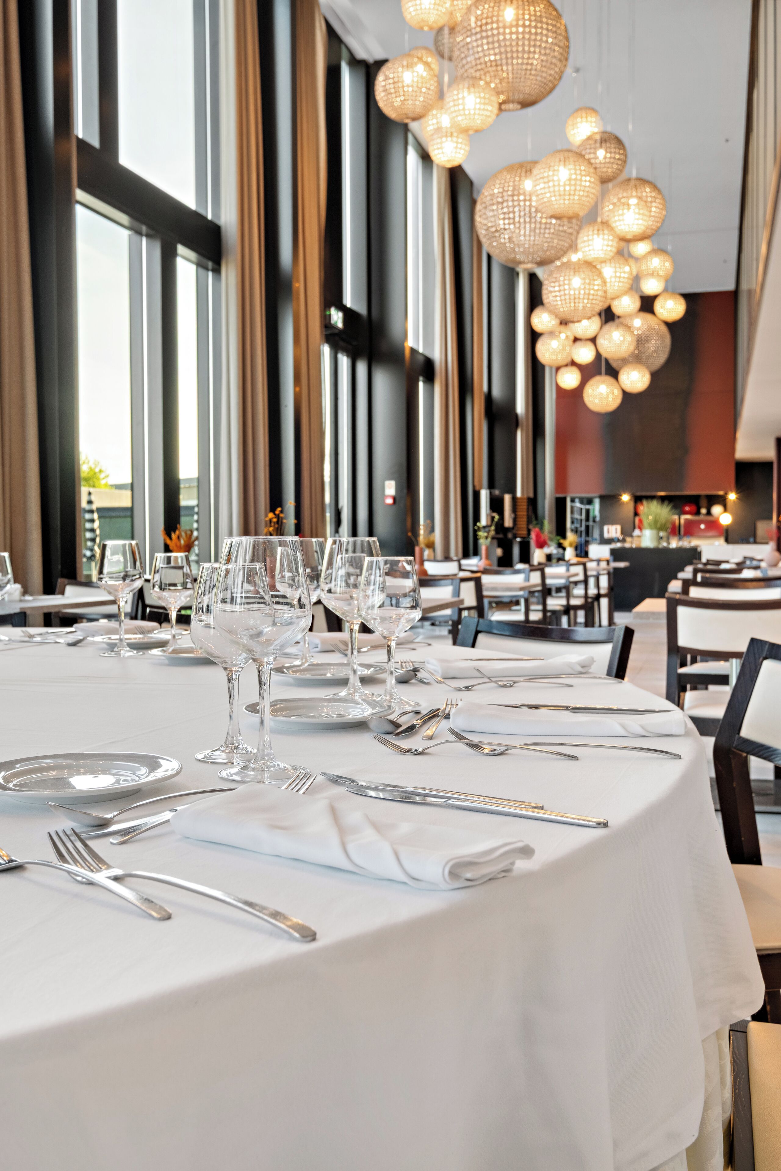 A restaurant with round tables set for dining, large windows, and hanging spherical light fixtures.