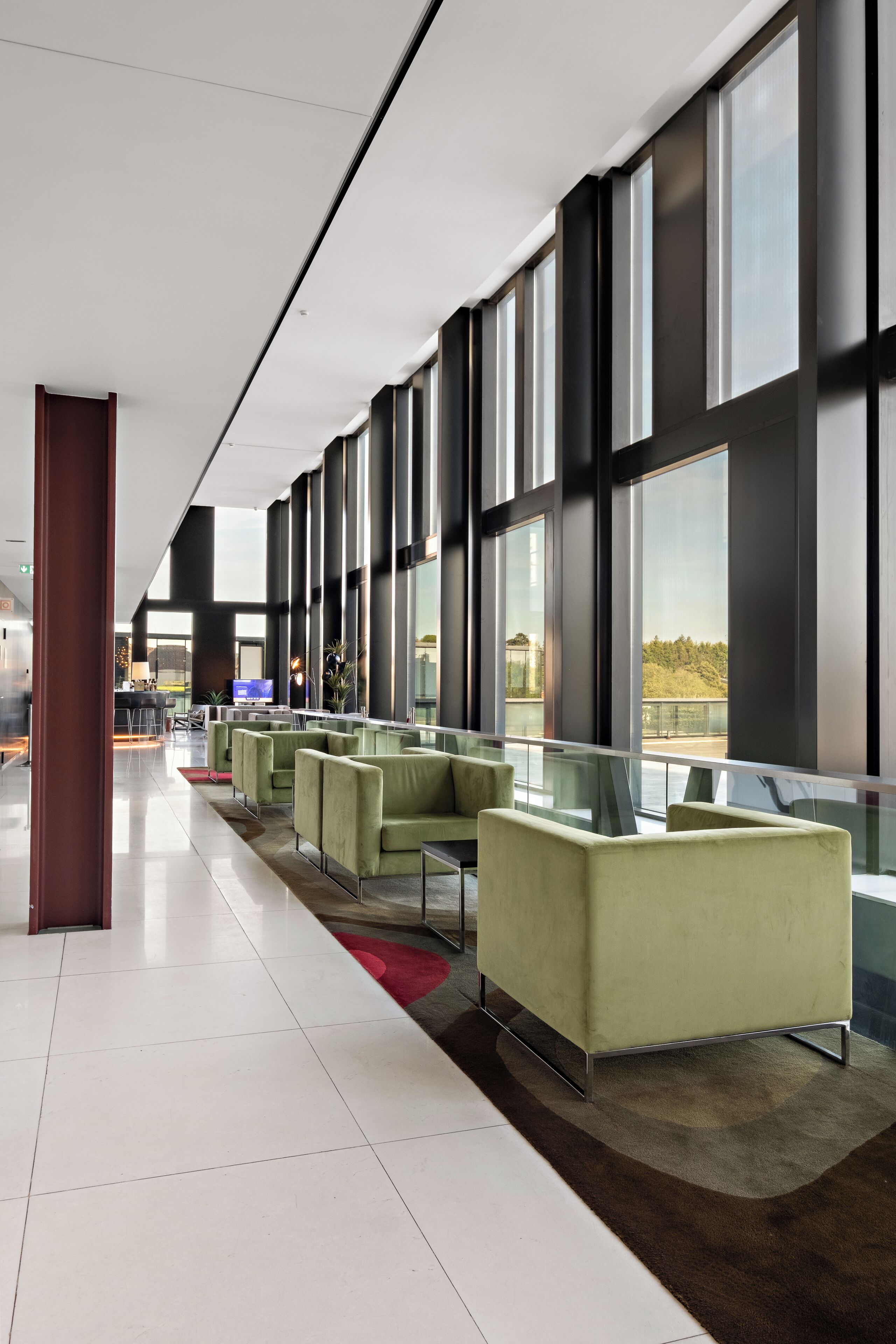 A modern hotel lobby with a long reception desk, green seating area, large windows, and a view of the outdoors.