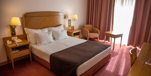 A hotel room with a large bed, two bedside tables with lamps, a chair, a desk, and a window with curtains.
