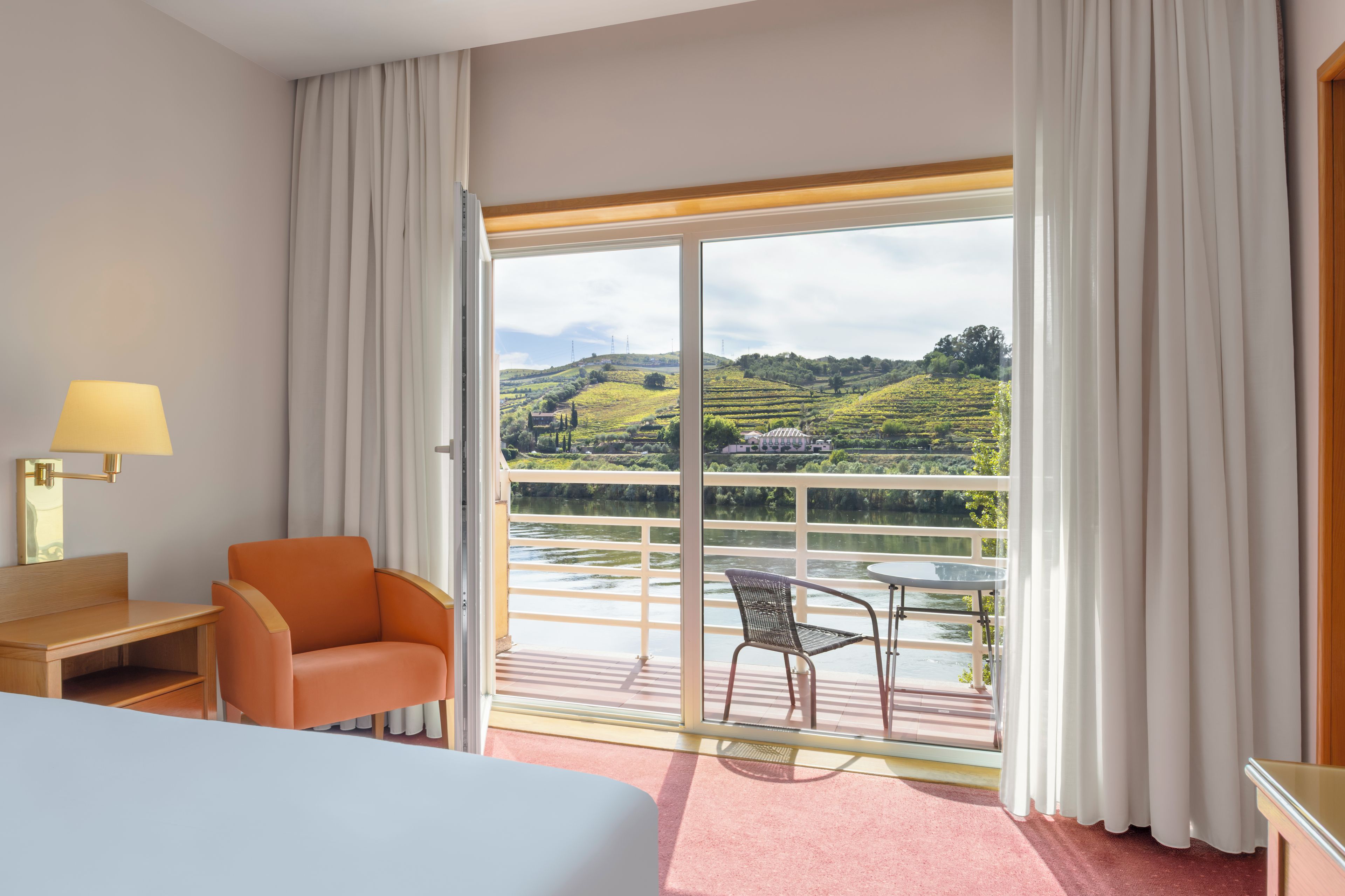 Habitación de hotel con cama, escritorio, silla, mesa y balcón con vistas al campo.