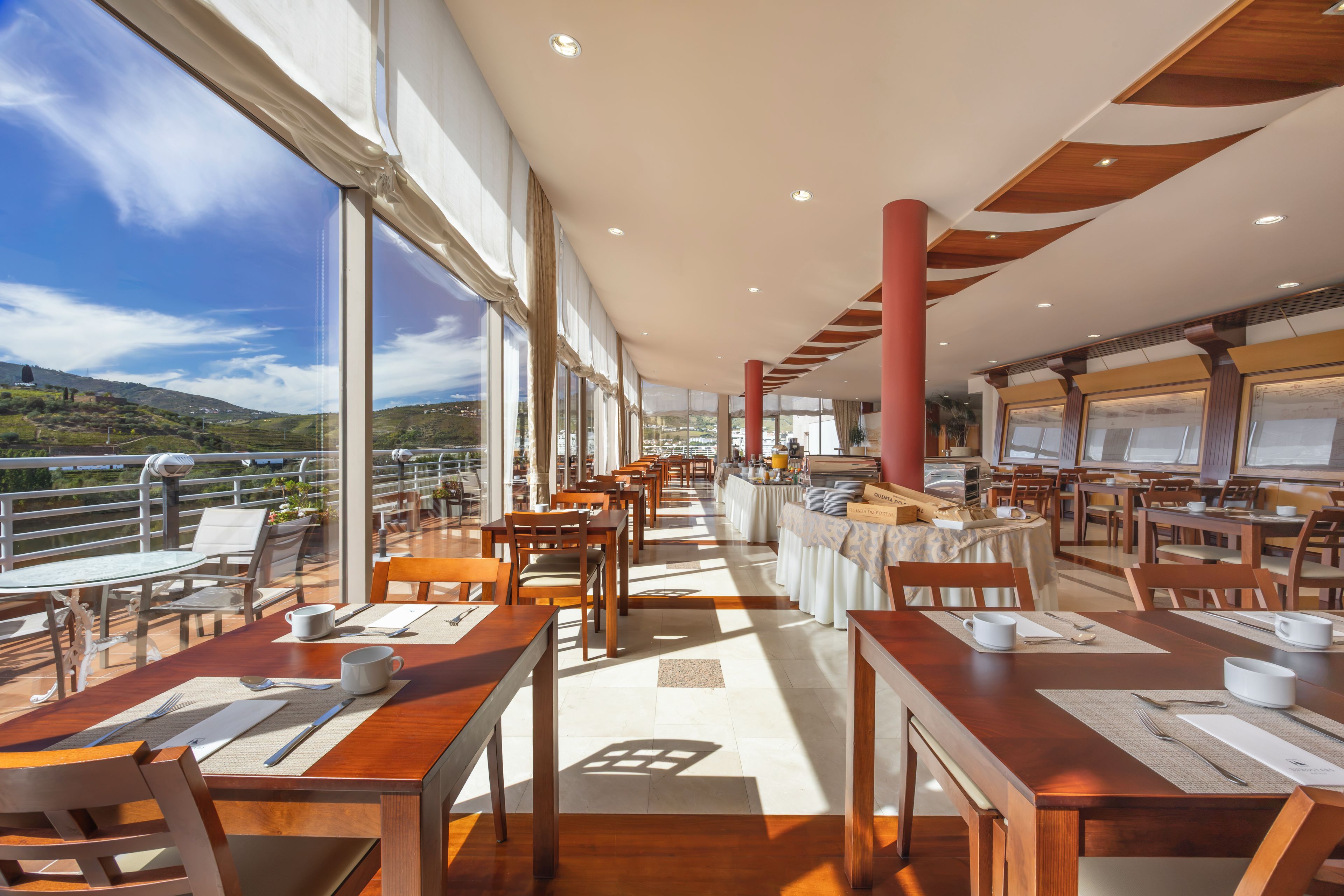 A spacious dining area with multiple tables and chairs, large windows offering a view of the landscape, and a modern ceiling design.