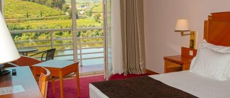 A hotel room with a bed, a desk, a chair, a nightstand, and a view of a green landscape through a large window.