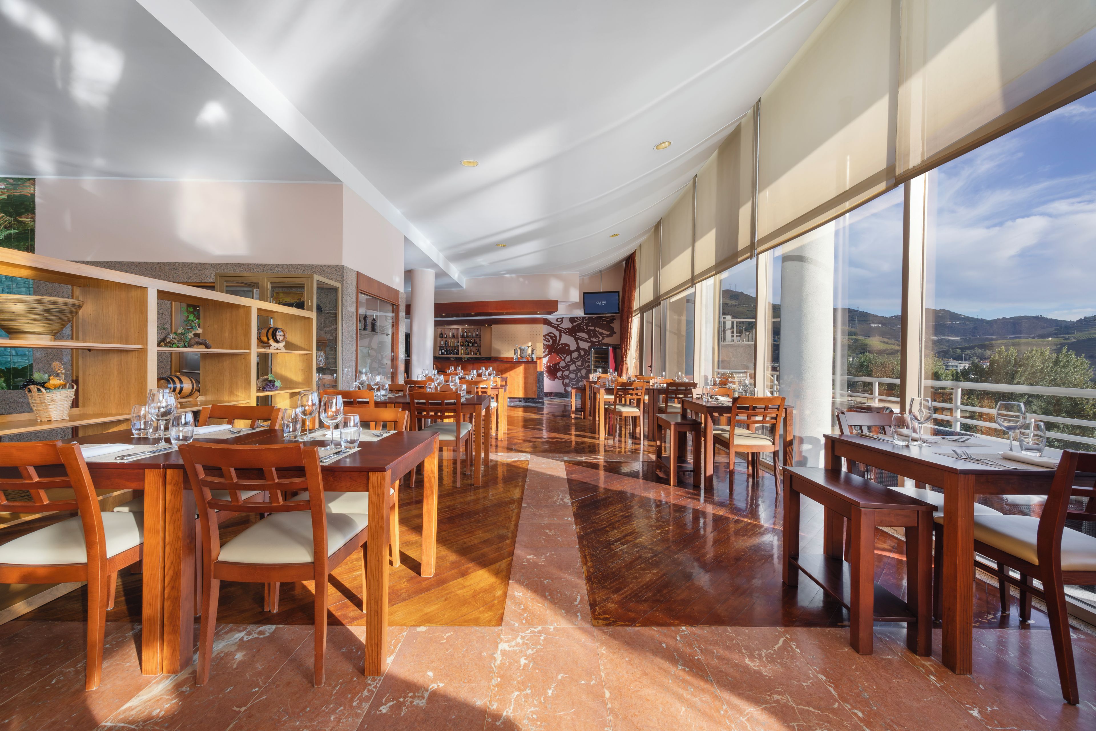 A spacious restaurant with multiple dining tables and chairs, a bar area, and large windows offering a view of the countryside.