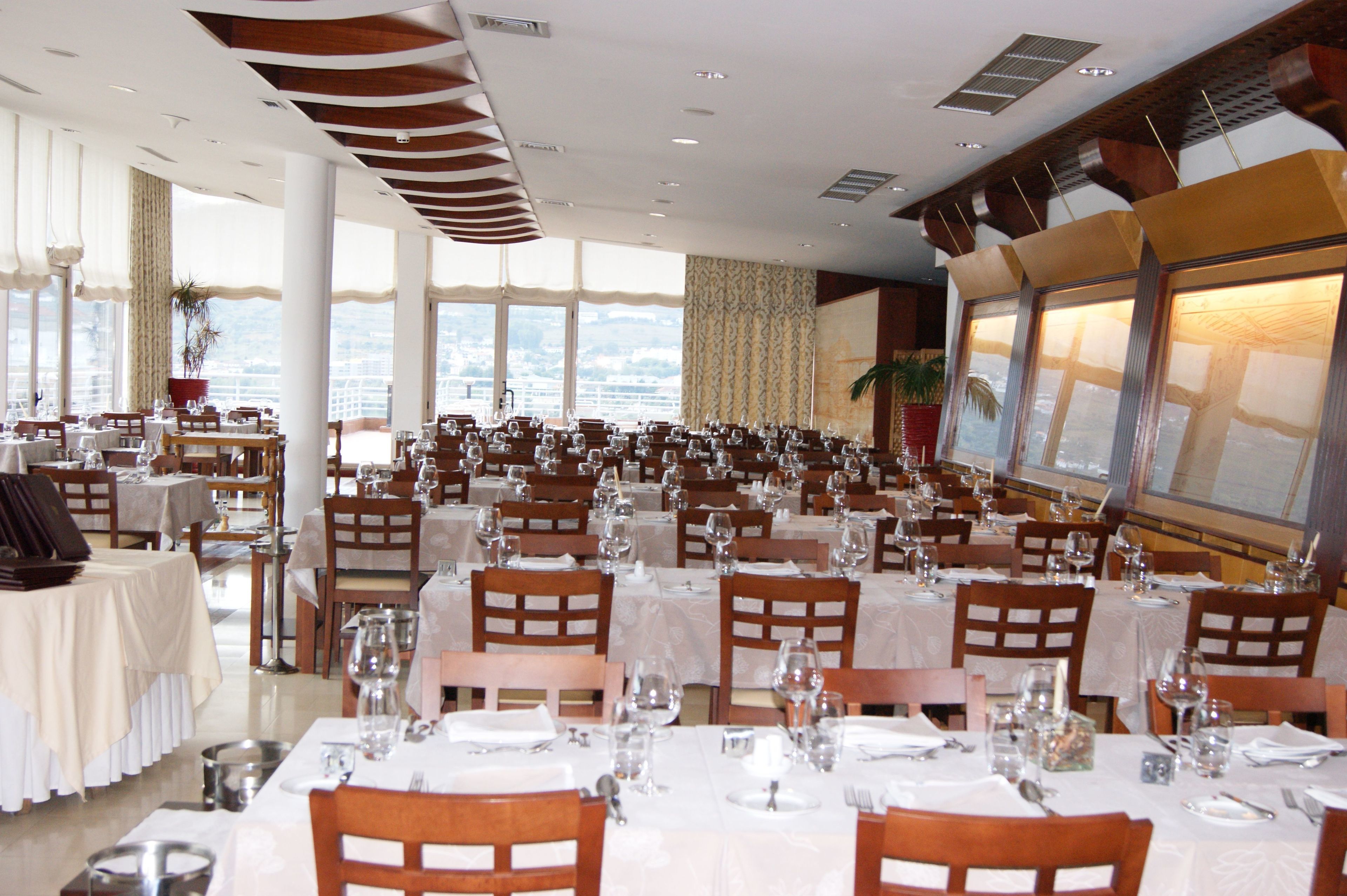 A large banquet hall with multiple tables set for dining, large windows offering a view, and a high ceiling with modern lighting fixtures.
