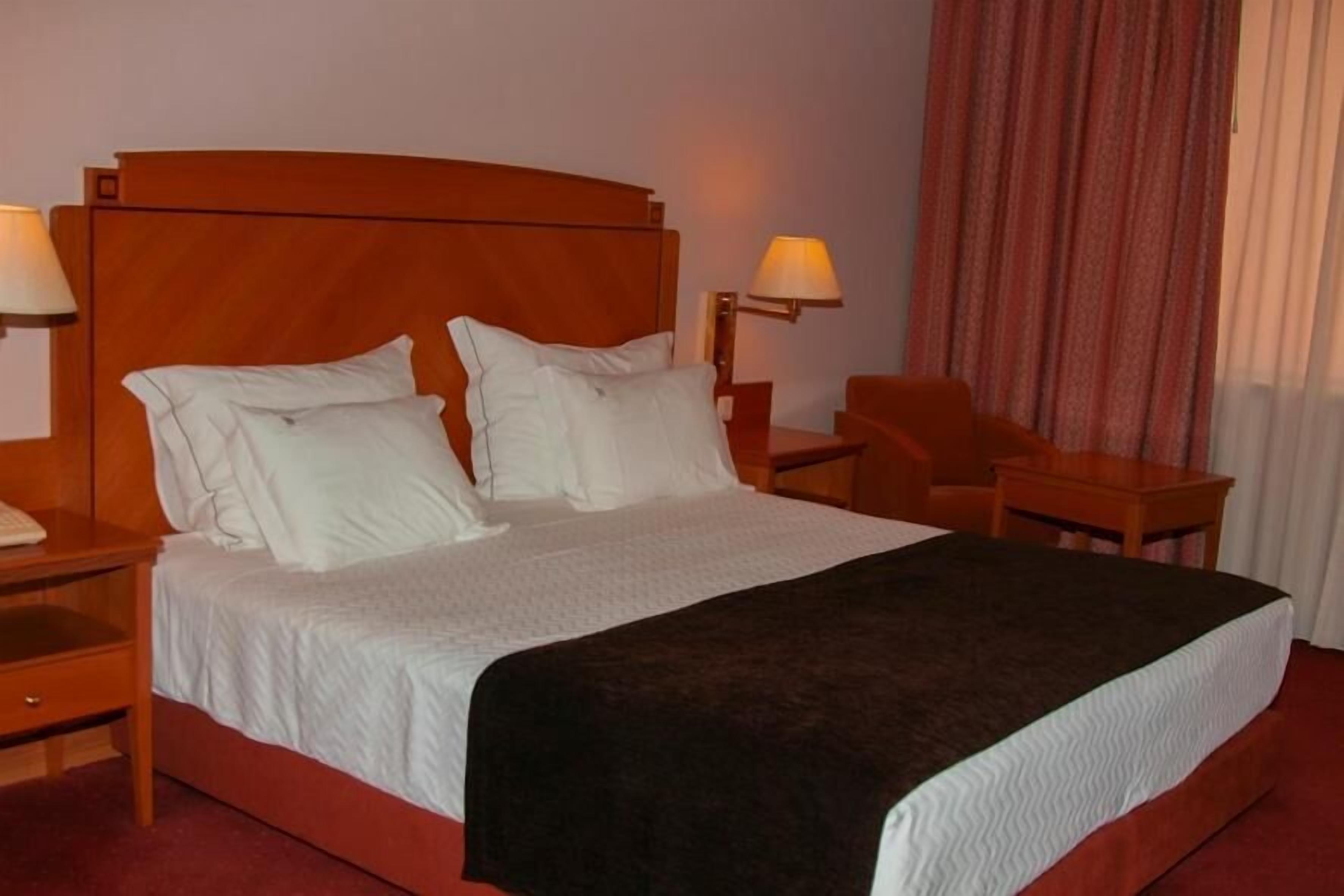 Una habitación de hotel con una cama grande, dos mesitas de noche, una silla y una lámpara.