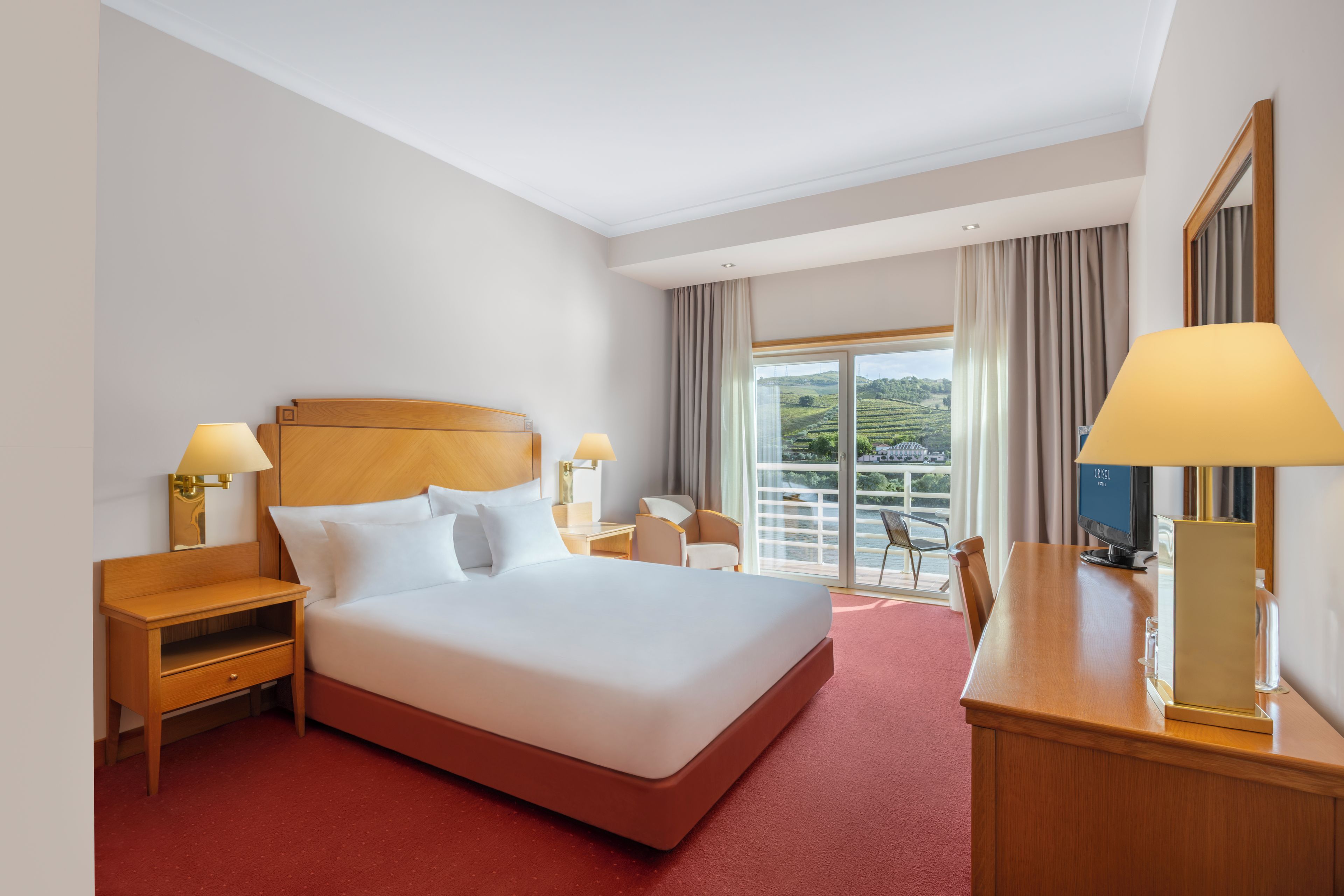 Une chambre d’hôtel avec un grand lit, deux tables de chevet, un bureau avec une chaise, une grande fenêtre avec des rideaux et une vue sur un paysage.