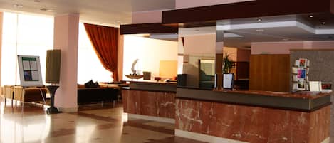 Zona de recepción de un hotel con mostrador de mármol, asientos y un amplio ventanal con cortinas.