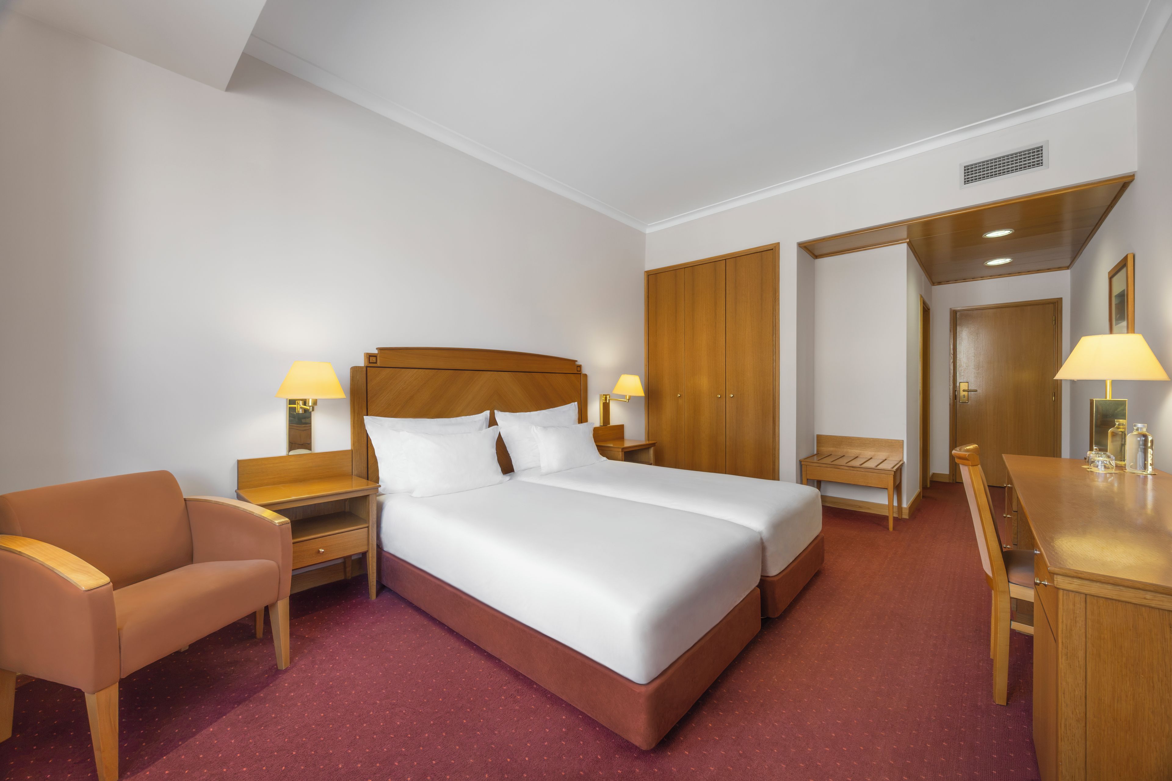 Habitación de hotel con una cama grande, dos mesitas de noche con lámparas, un armario de madera, un escritorio con una silla y un pequeño banco.