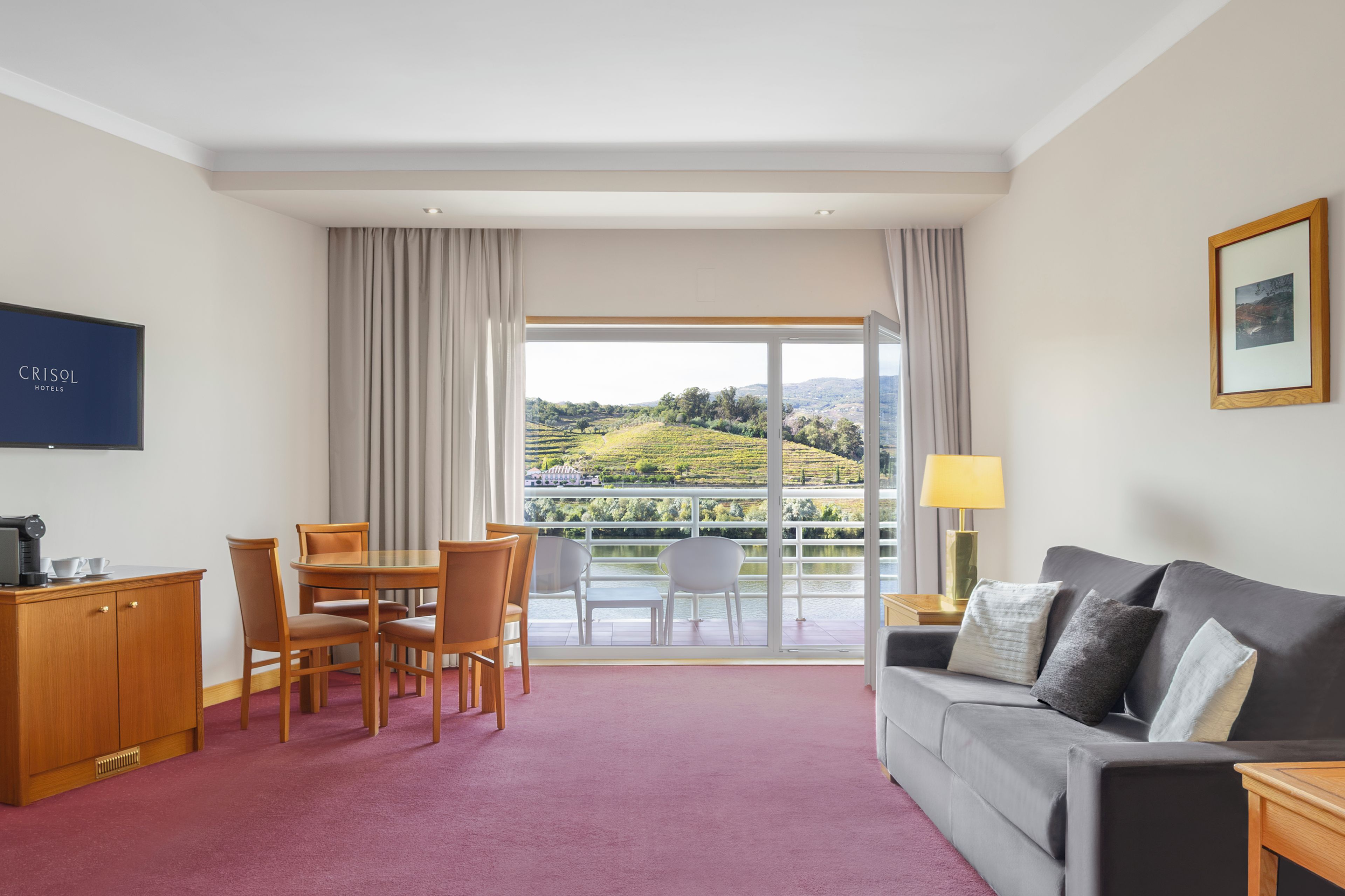 A hotel room with a balcony, a dining area, a sofa, and a TV.