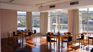 A spacious room with a large dining table and chairs, large windows with curtains, and a view of a river and hills.