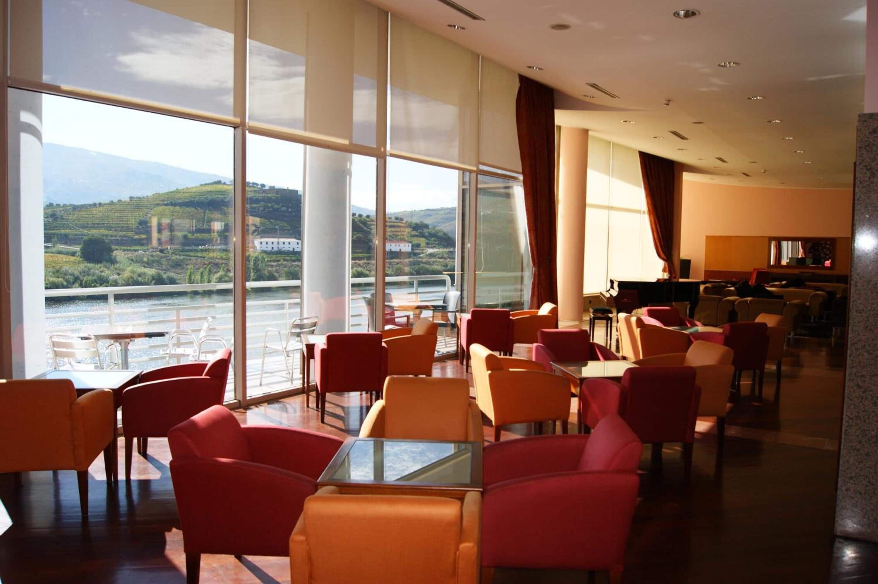 Une salle à manger d’hôtel dotée de grandes fenêtres, de chaises rouges et orange, et offrant une vue sur une rivière et des collines.