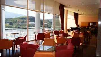 Une salle à manger d’hôtel dotée de grandes fenêtres, de chaises rouges et orange, et offrant une vue sur une rivière et des collines.