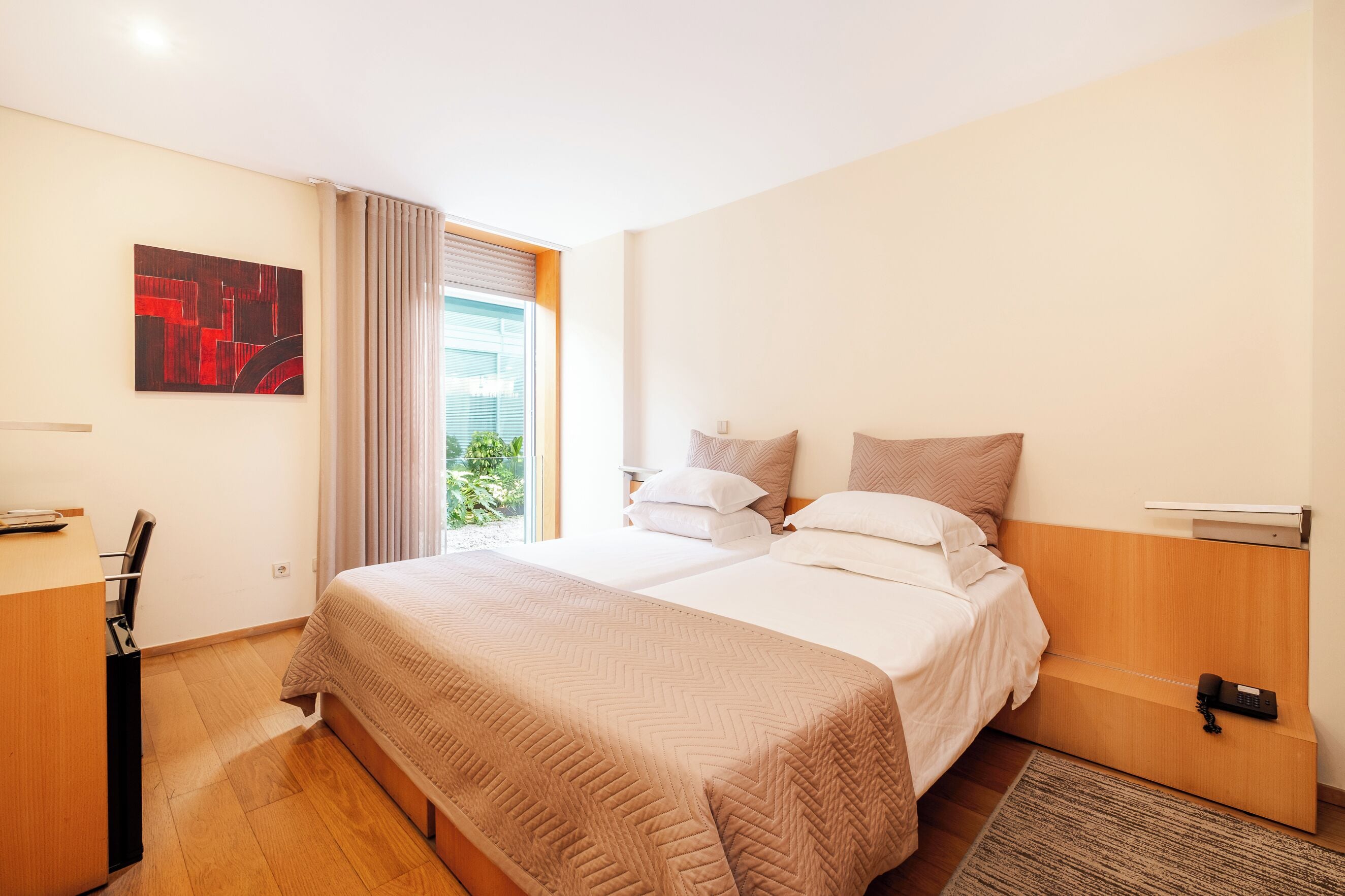 Hotellihuone, jossa on sänky, työpöytä, tuoli, ikkuna verhoilla ja taulu seinällä.