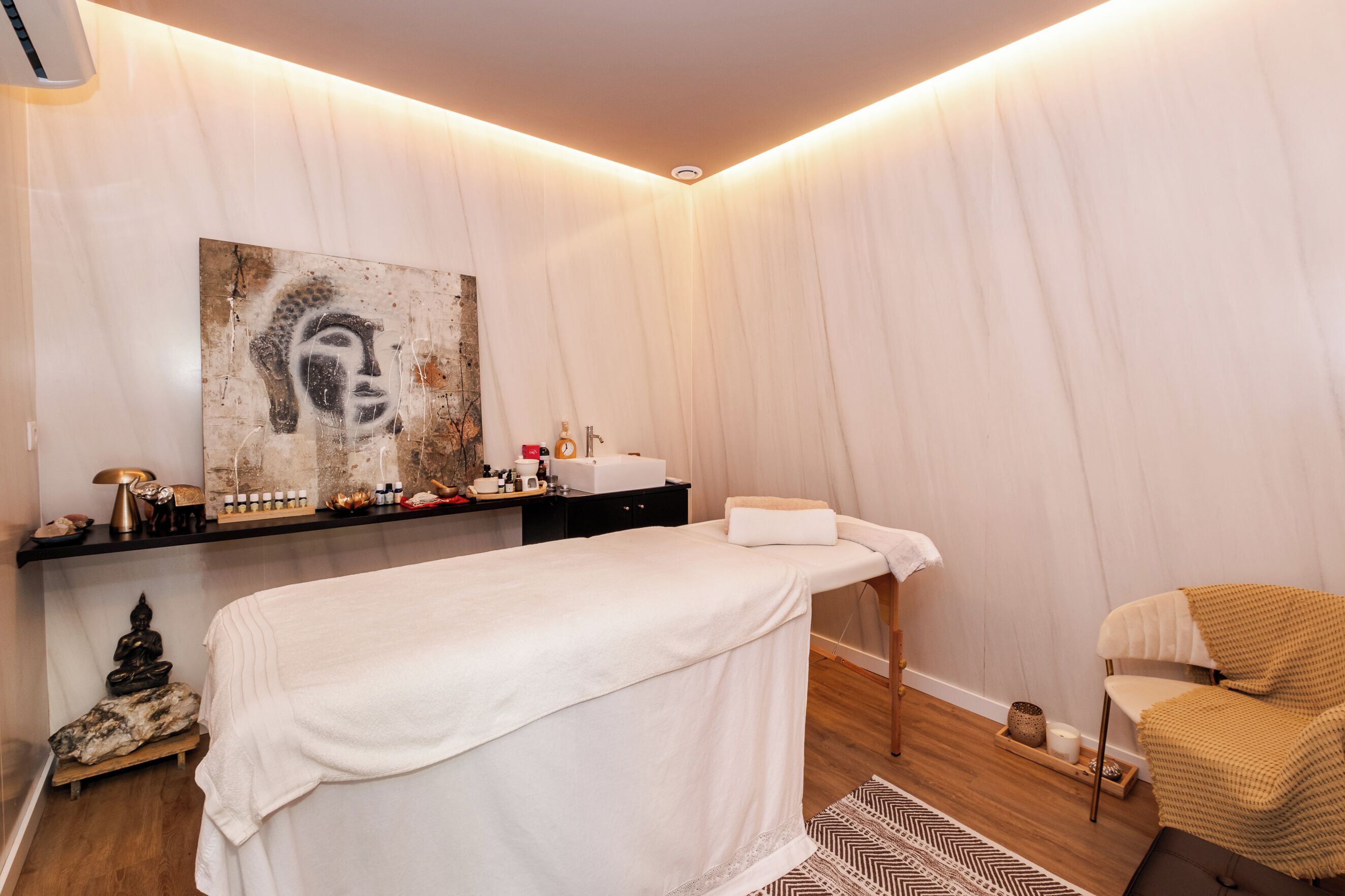 Ein moderner Spa-Raum mit Massagetisch, Buddha-Statue, einer an der Wand montierten Ablage mit Toilettenartikeln und einem großen Buddha-Gemälde.