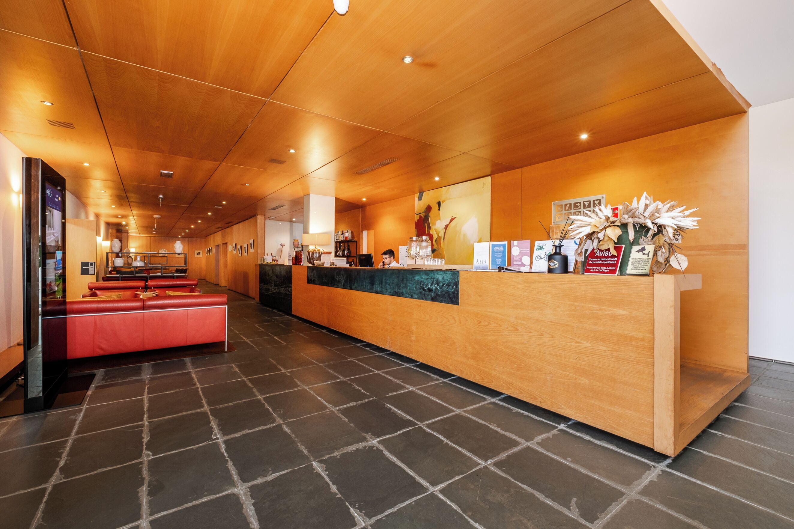 A hotel reception area with a wooden reception desk, a red seating area, and a tiled floor.