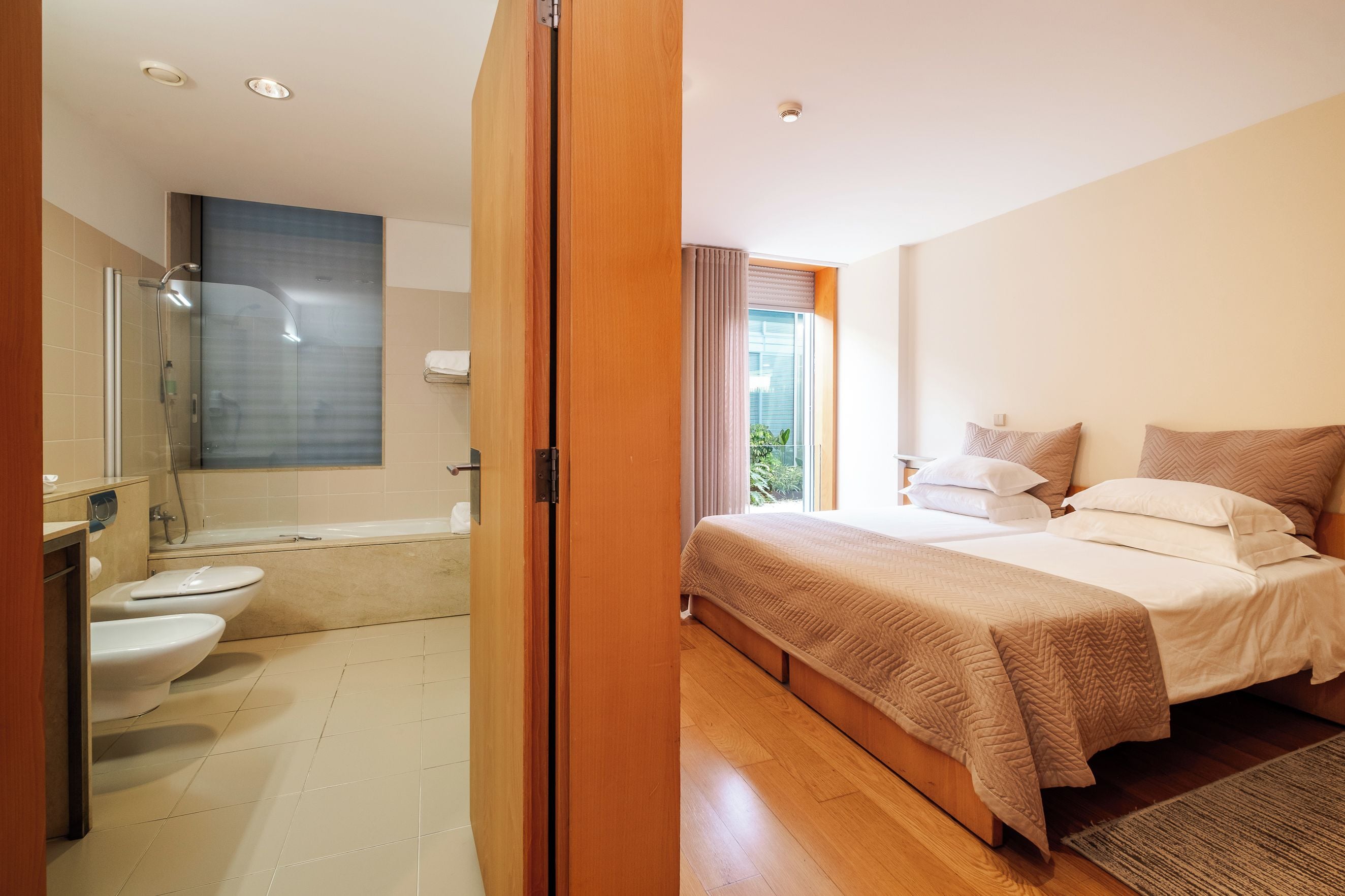 Een moderne badkamer met een glazen douchecabine, een badkuip en een toilet. De slaapkamer heeft een groot bed met een beige sprei en twee kussens.